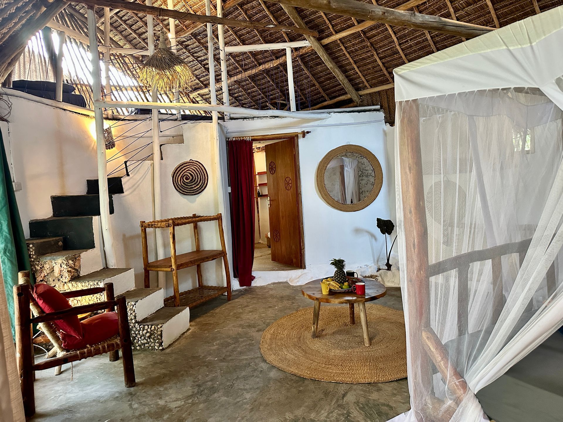 Cozy interior: white walls, bed with mosquito net, red chair, wooden furniture, round rug, stairs, door to another room.