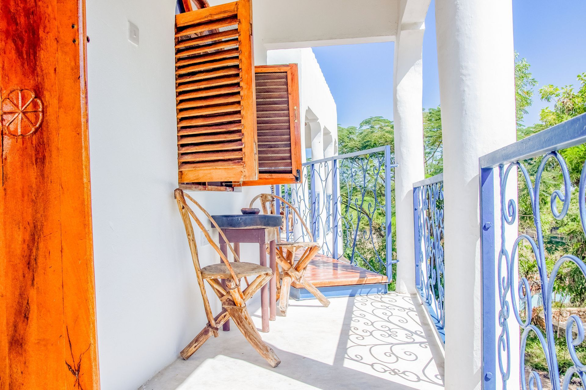 Balcony with wooden chairs and table, blue railing, wooden shutters, white walls, and bright sunlight.
