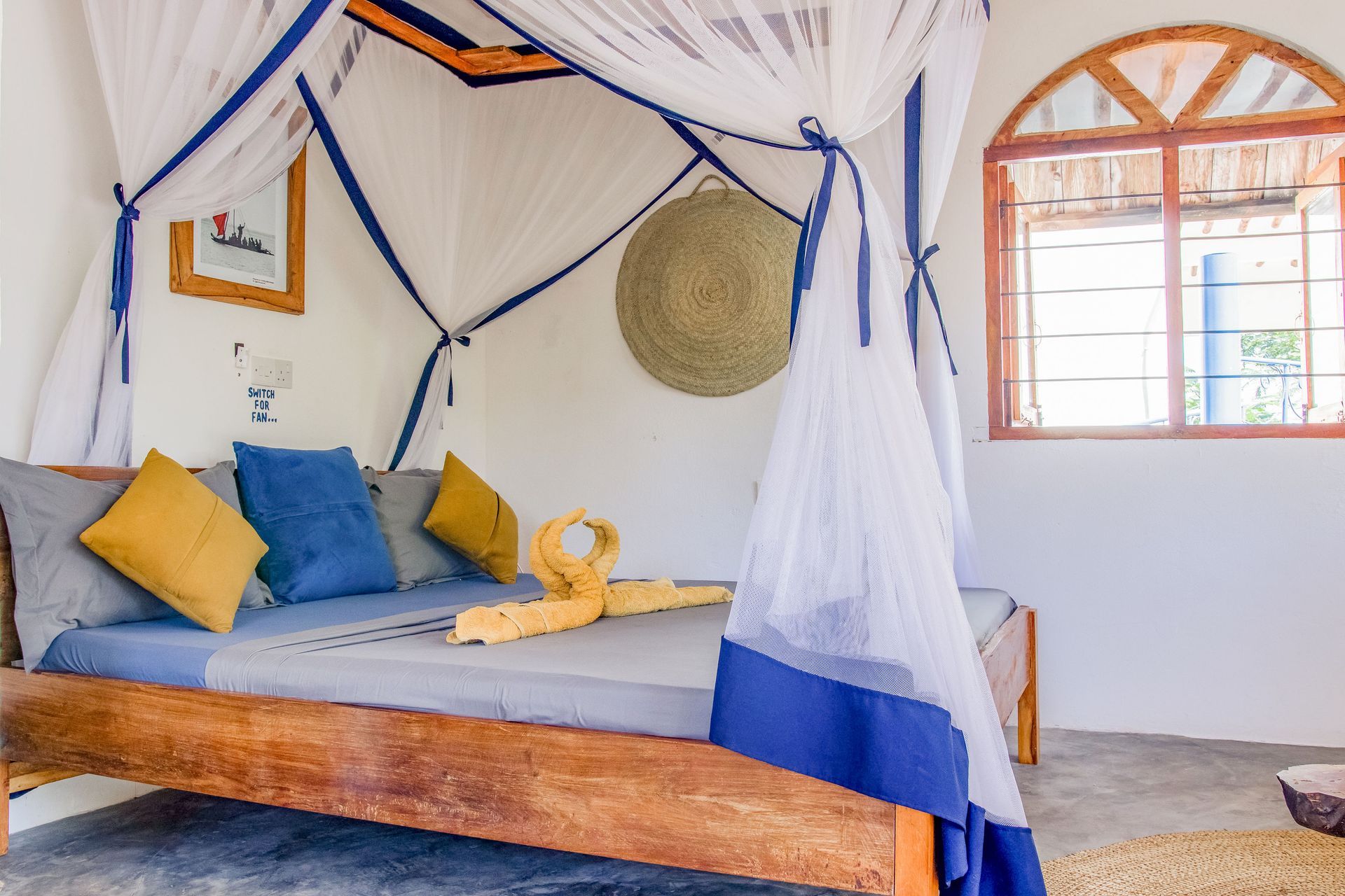 Bedroom with canopy bed; blue and yellow pillows; woven wall decor; window.