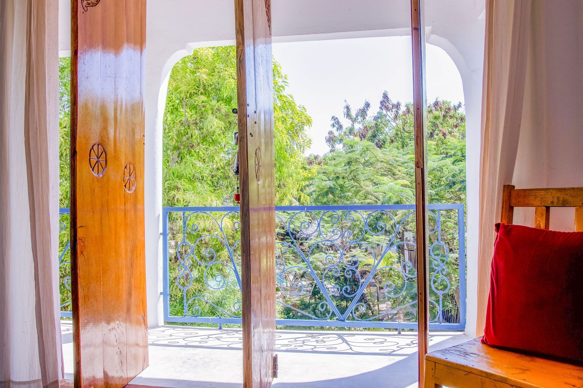 Open wooden doors reveal a balcony with a green tree view, red pillow on a wooden bench.