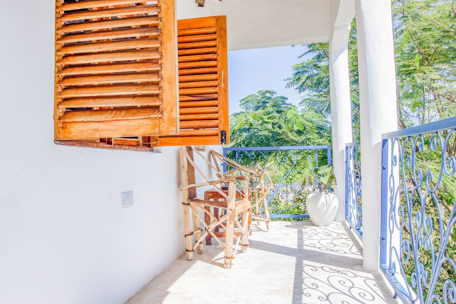 Balcony with wooden chairs, shutters, and ornate blue railing, overlooking green trees on a sunny day.