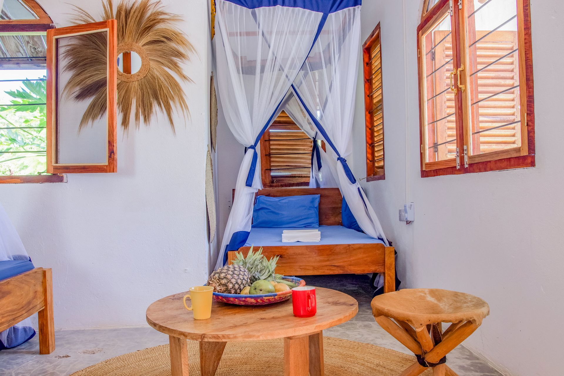 Cozy room with bed under mosquito net, wooden furniture, fruit on table, windows with natural light.