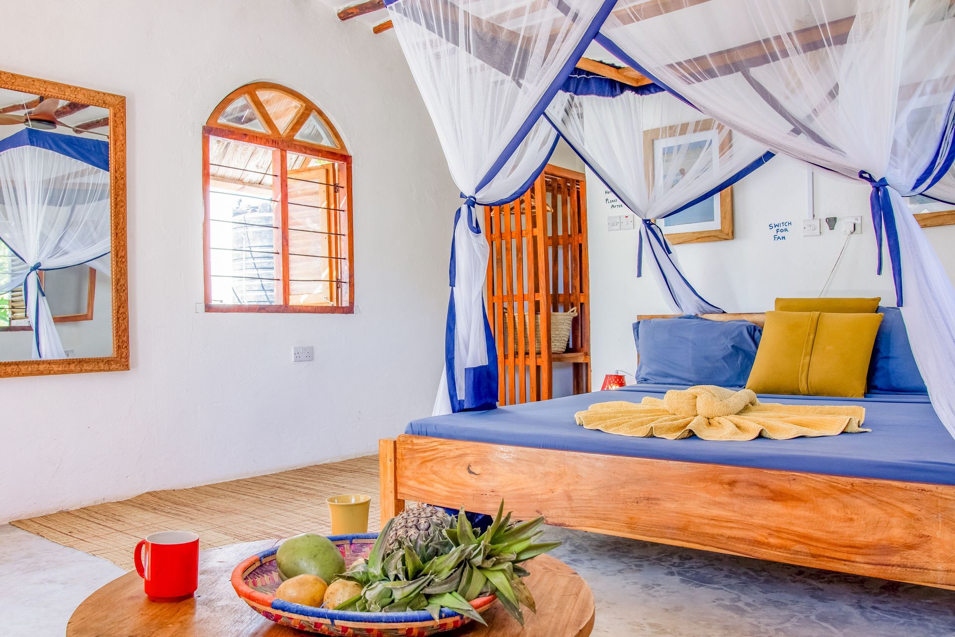 Bedroom with canopy bed, fruit basket, and mirror. Bright white walls, wooden accents, and blue bedding.