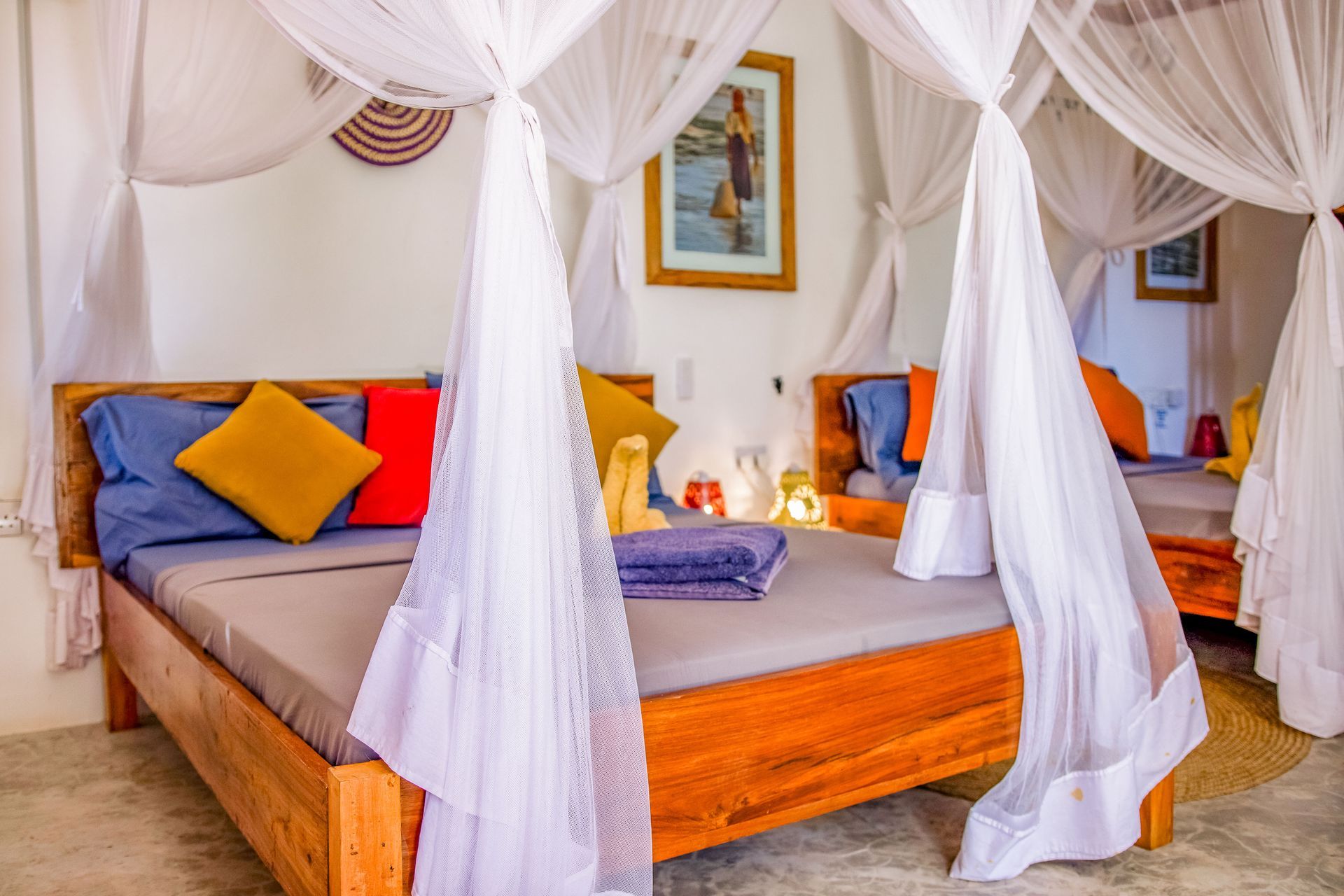 Bedroom with wooden bed frame, mosquito net, colorful pillows, and artwork on the wall.