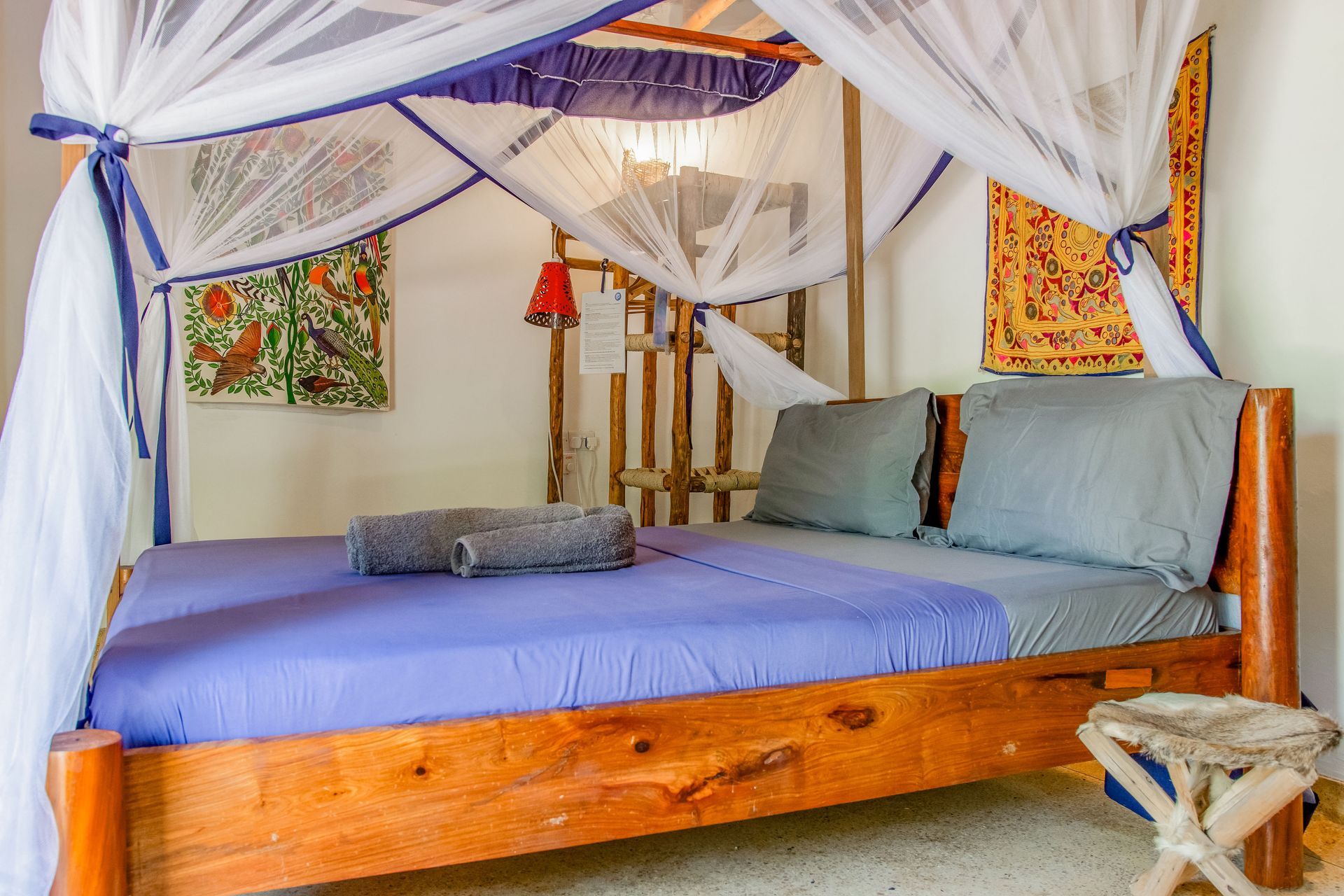 Bedroom with wooden bed, mosquito net, art, blue and gray bedding, and a small stool.