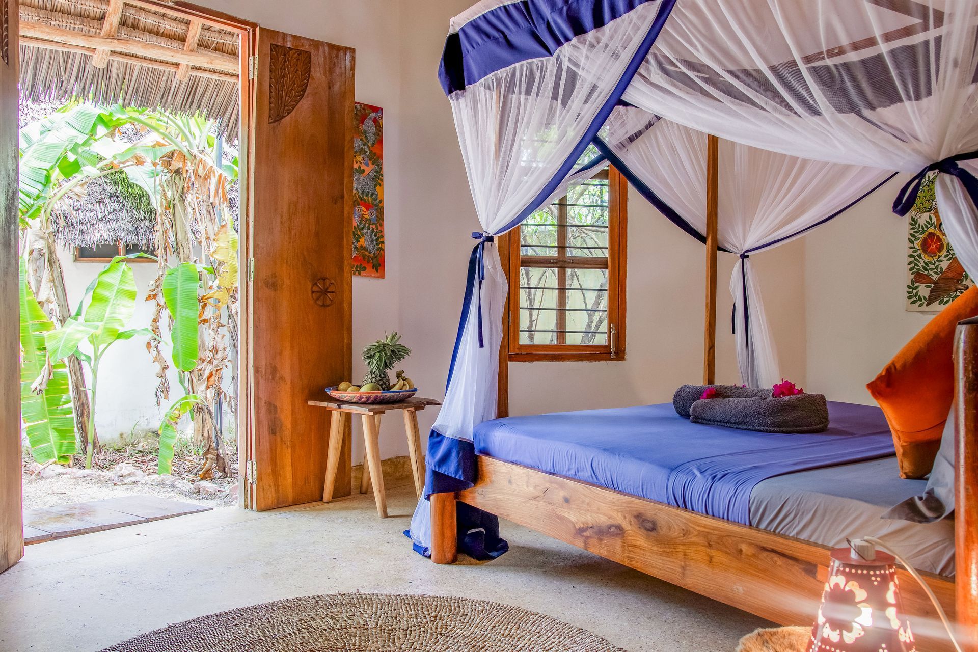 Bedroom with canopy bed, wooden door open to greenery, rug on the floor, white walls.
