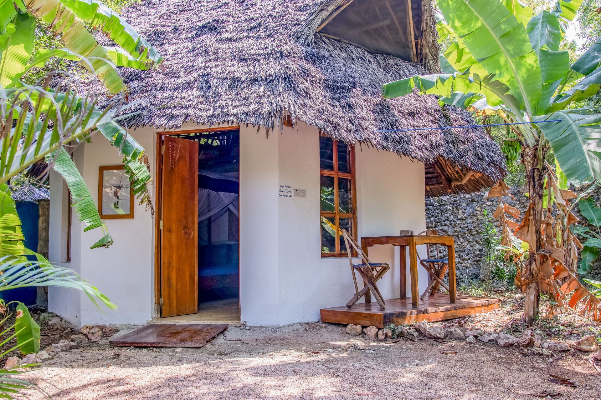 White cottage with thatched roof, wooden door, and small porch surrounded by lush greenery.