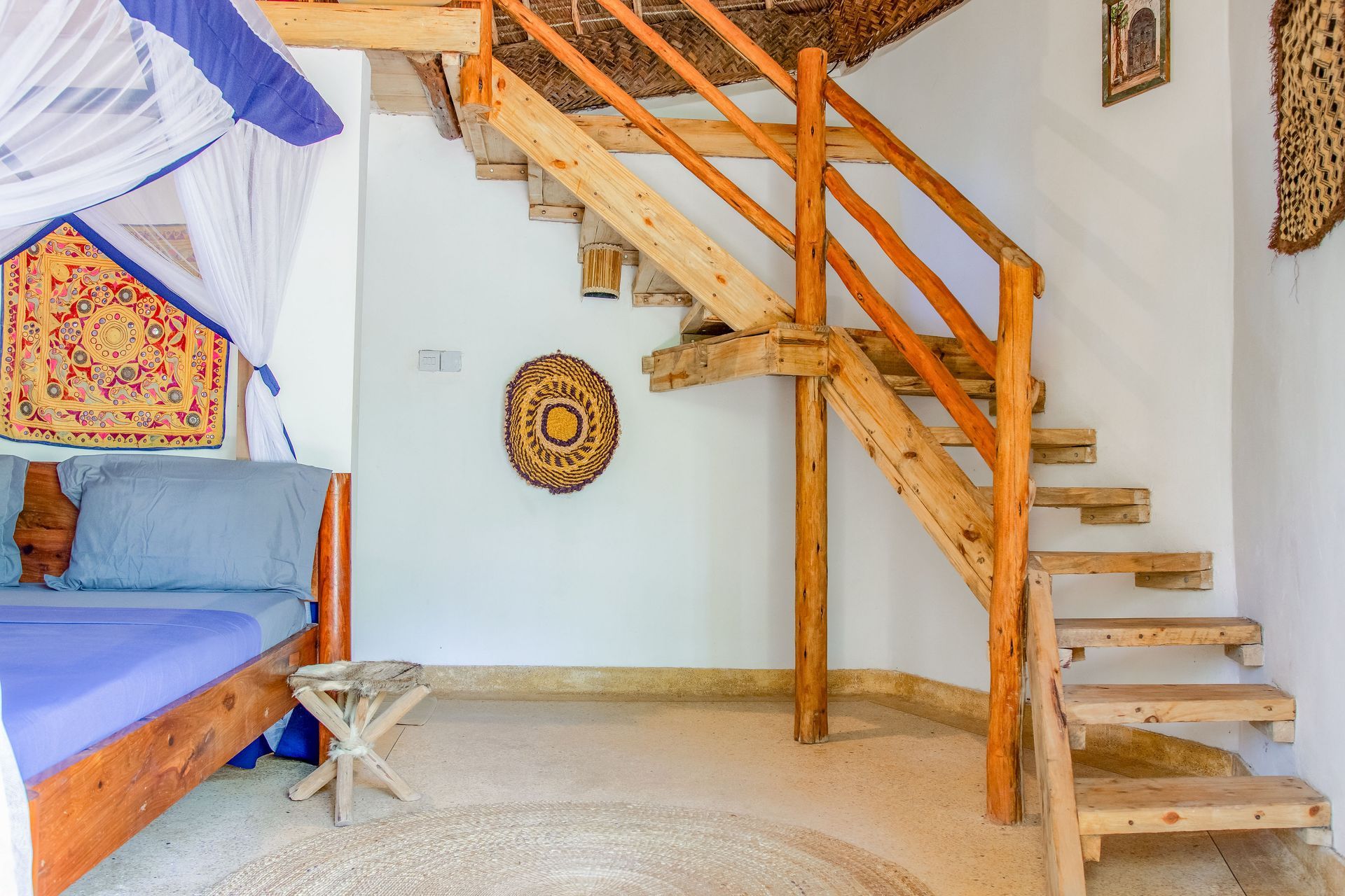 Wooden staircase in a room with a bed, woven wall decorations, and a thatched ceiling.