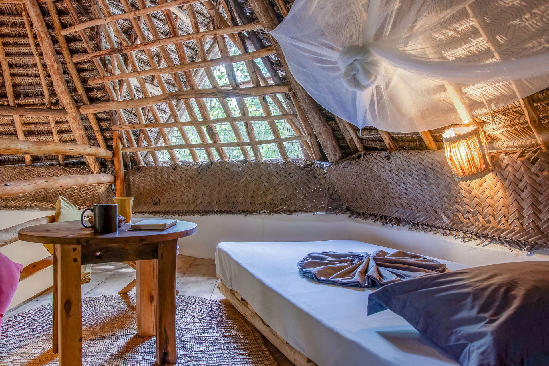 Cozy, thatched-roof bedroom with a bed, table, and mosquito net. Sunlight streams through the windows.