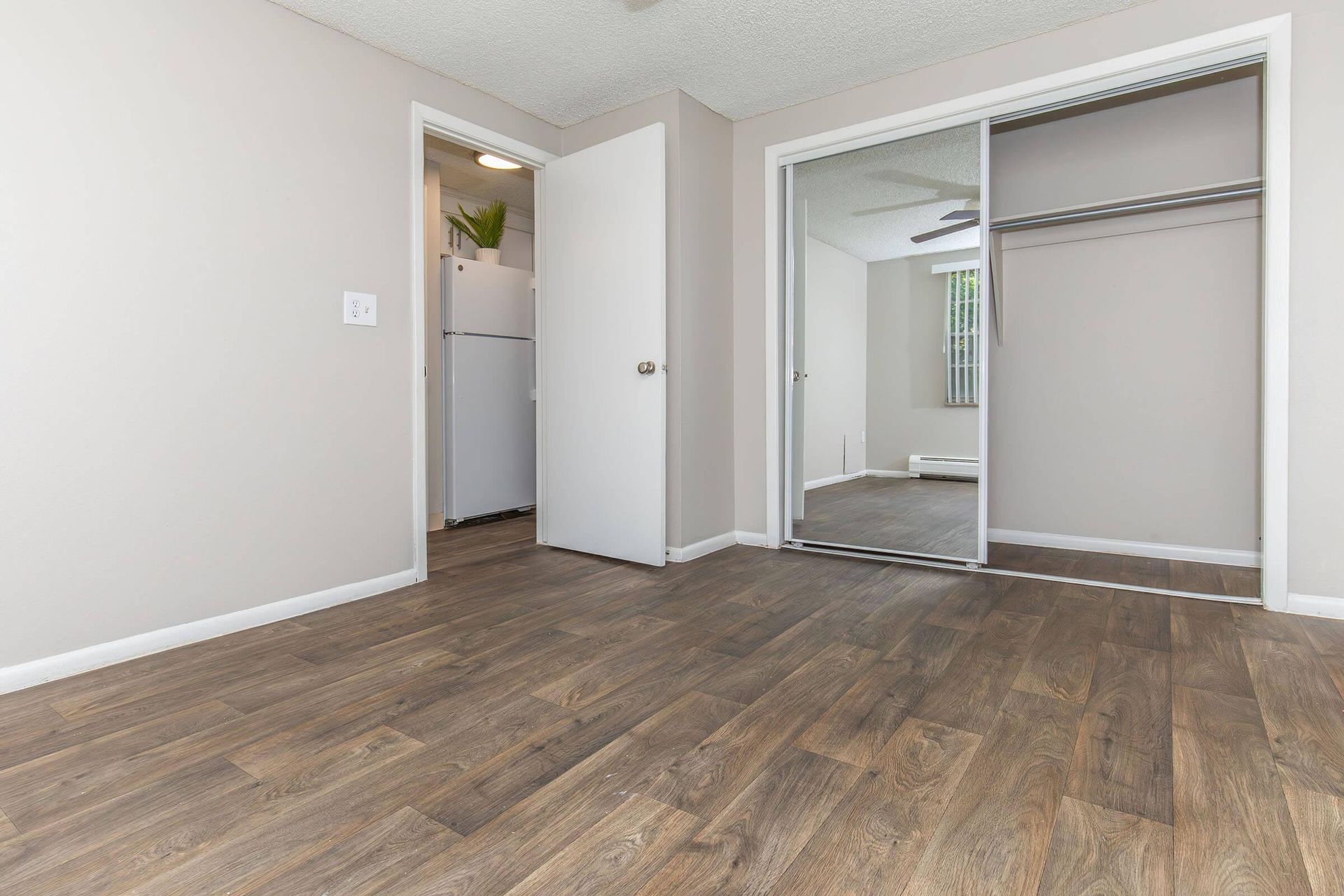 Empty bedroom with wood-look flooring, light gray walls, mirrored closet, and open door to a small kitchen.