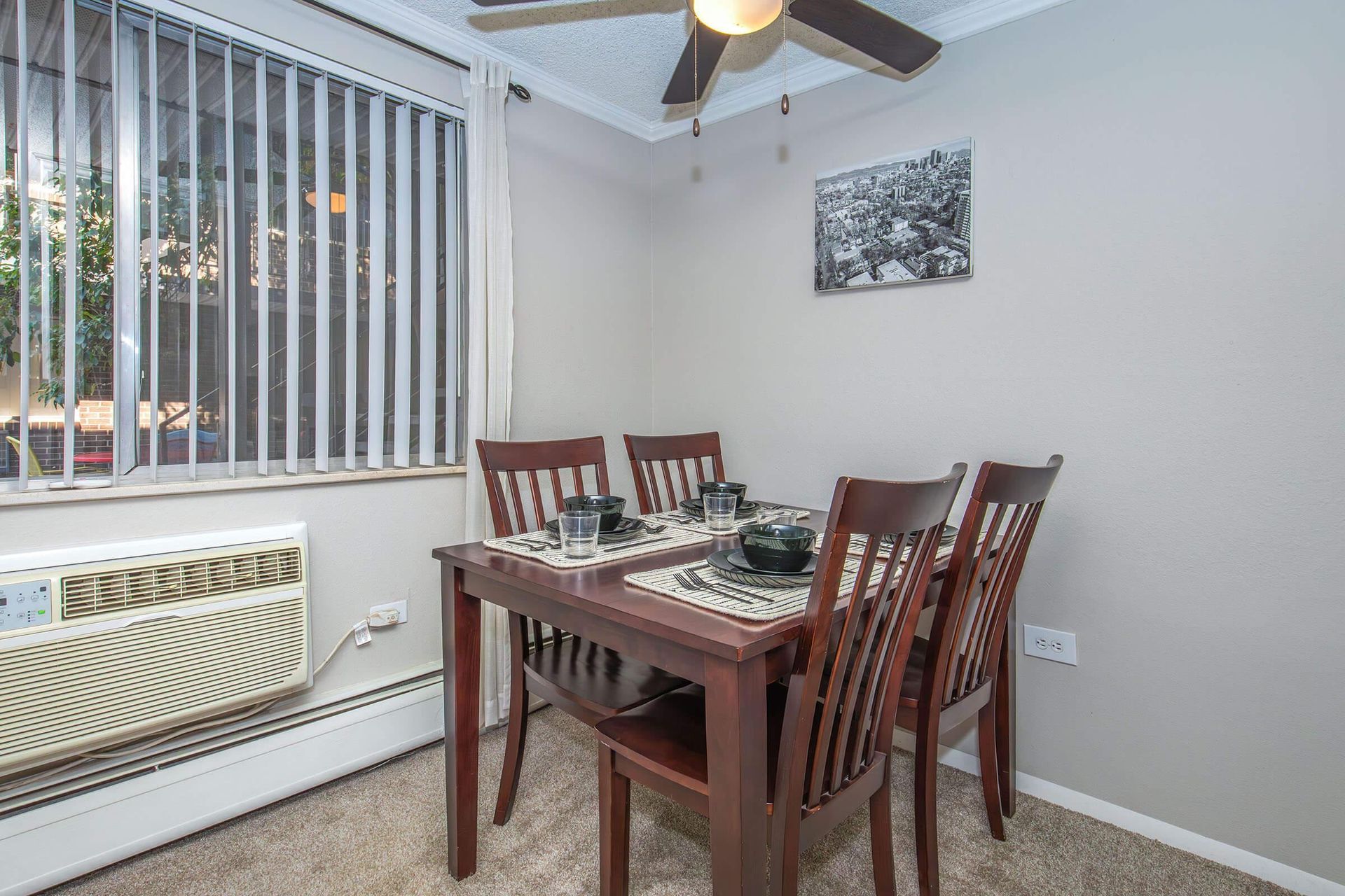 Dining area with wooden table set for four, window with blinds, and air conditioner.