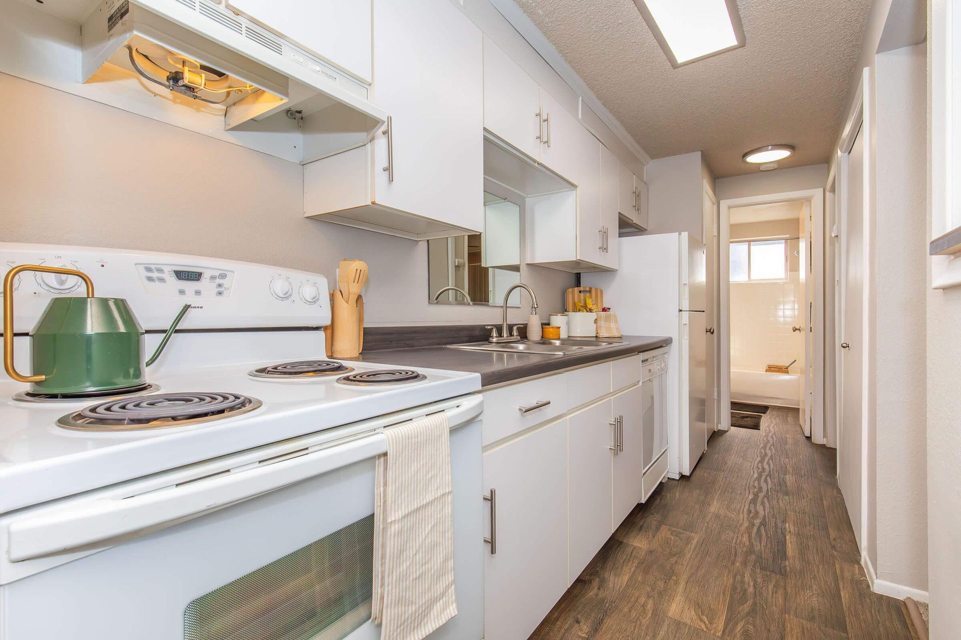 White kitchen with stove, cabinets, sink, and refrigerator; doorway to bathroom.