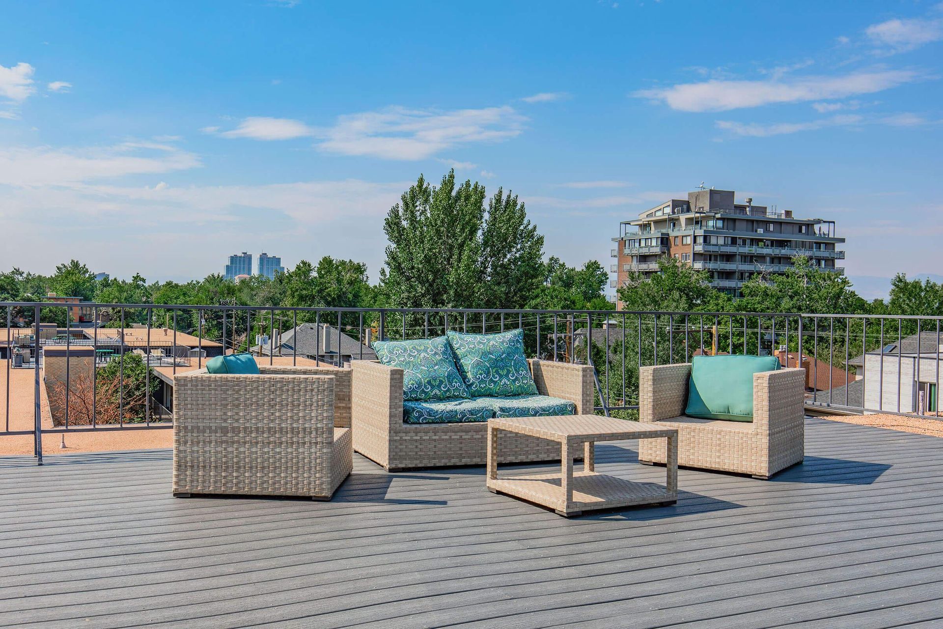 Rooftop patio with beige wicker furniture, turquoise cushions, and a city view on a sunny day.