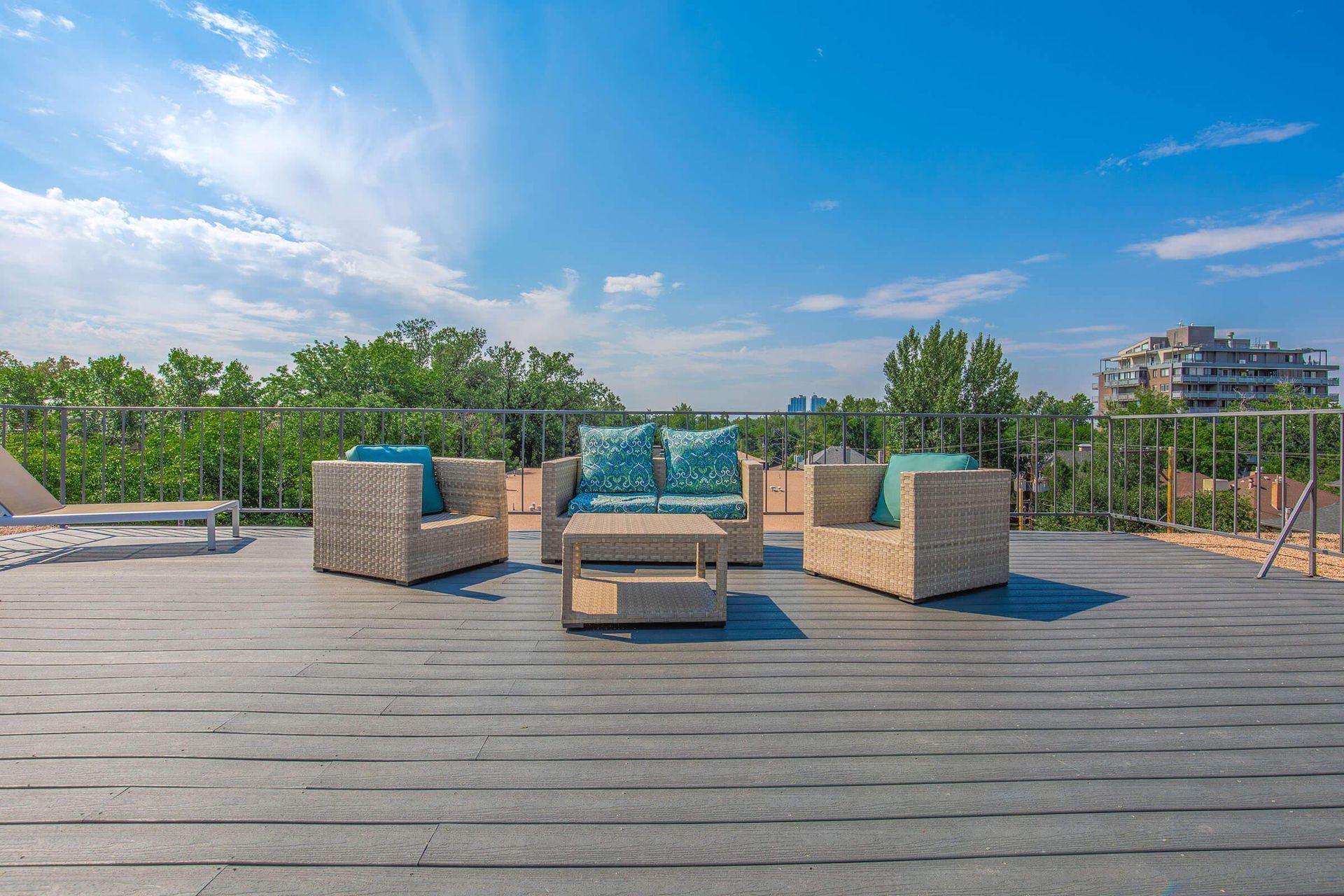 Rooftop patio with woven seating, teal cushions, and a small table, against a backdrop of trees and a bright blue sky.