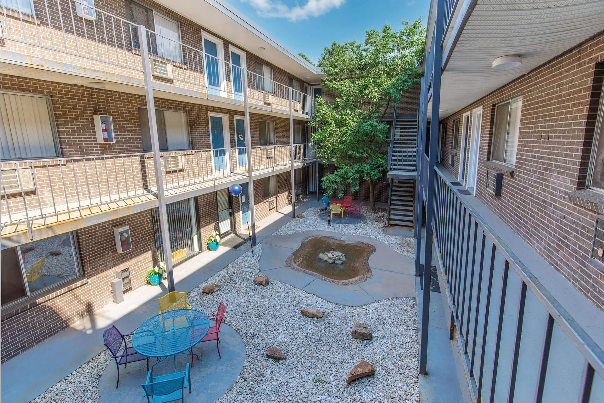 Apartment complex courtyard with brick buildings, balconies, and a pebble-filled area with a small water feature and seating.