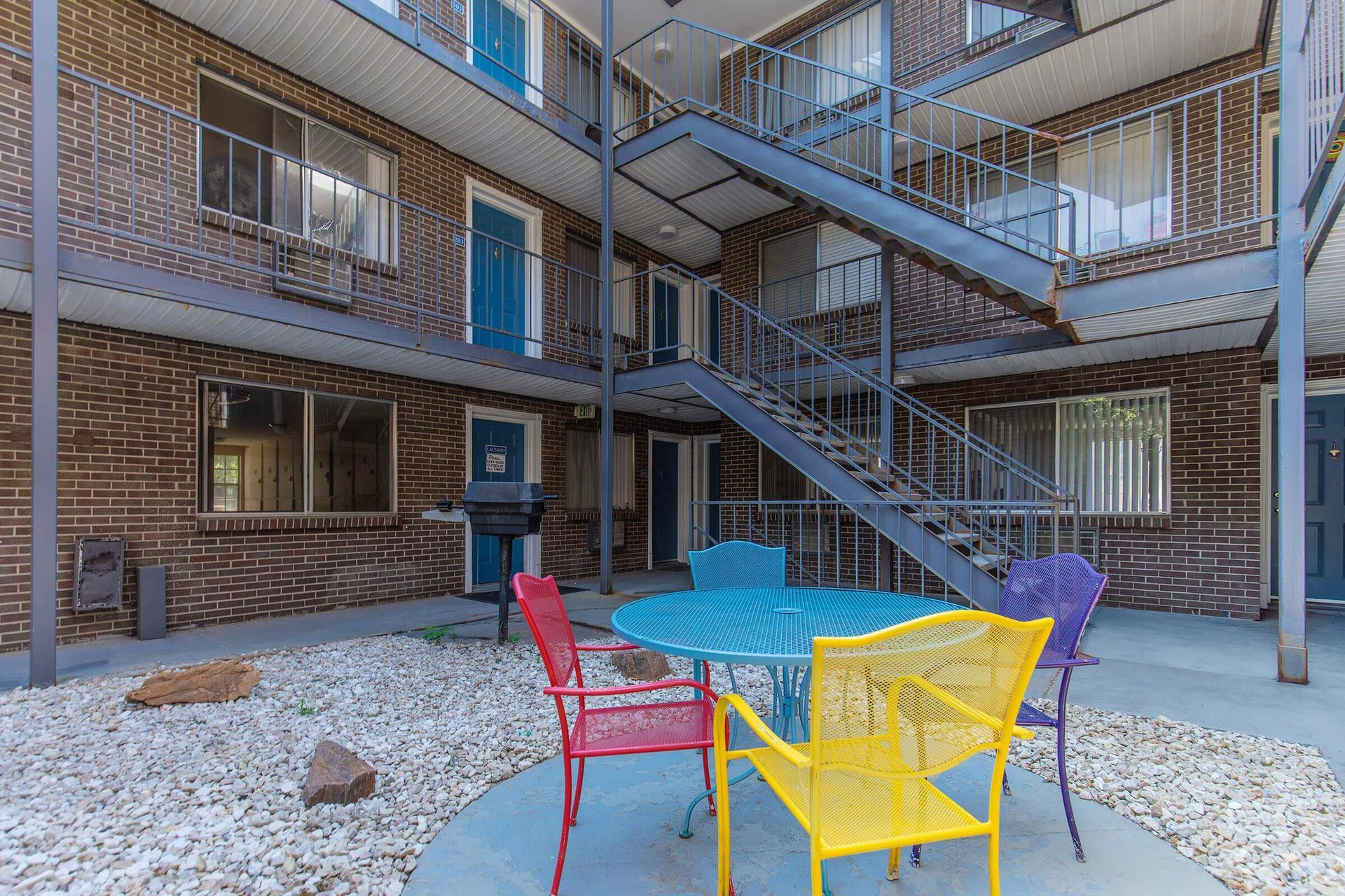 Courtyard with metal staircase, brick building, and colorful chairs around a table on a gravel surface.