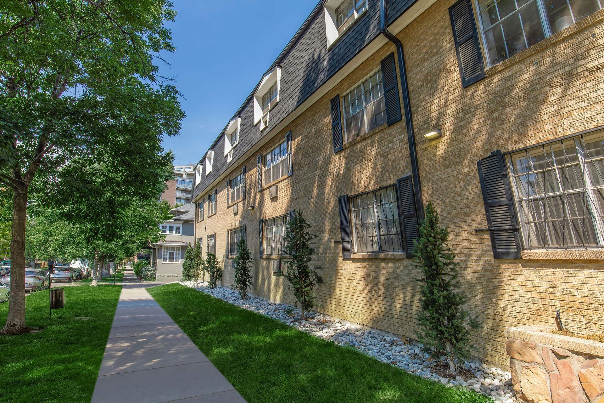 Apartment building with stone facade, black shutters, and black roof. Green lawn and sidewalk in front.