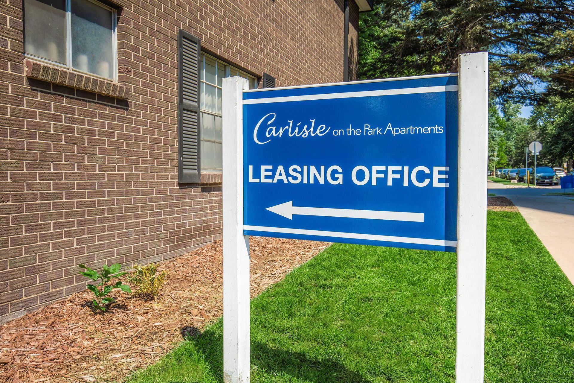 Sign for Carlisle on the Park Apartments Leasing Office with directional arrow, by a brick building and green lawn.