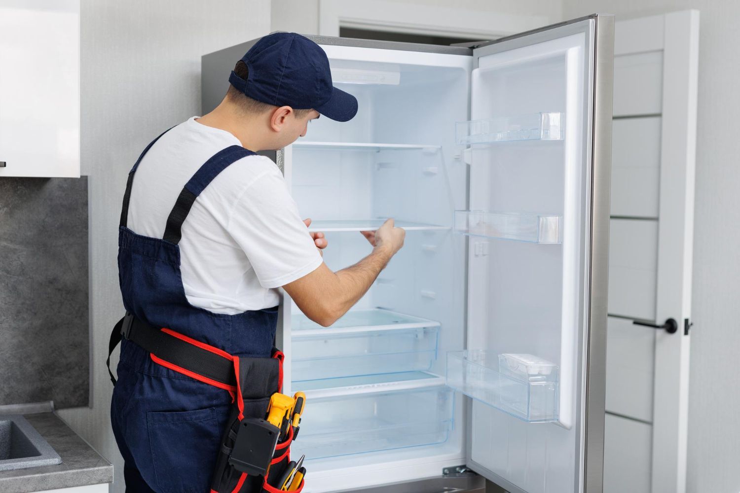 Technician in overalls inspecting an open refrigerator in a kitchen or workshop