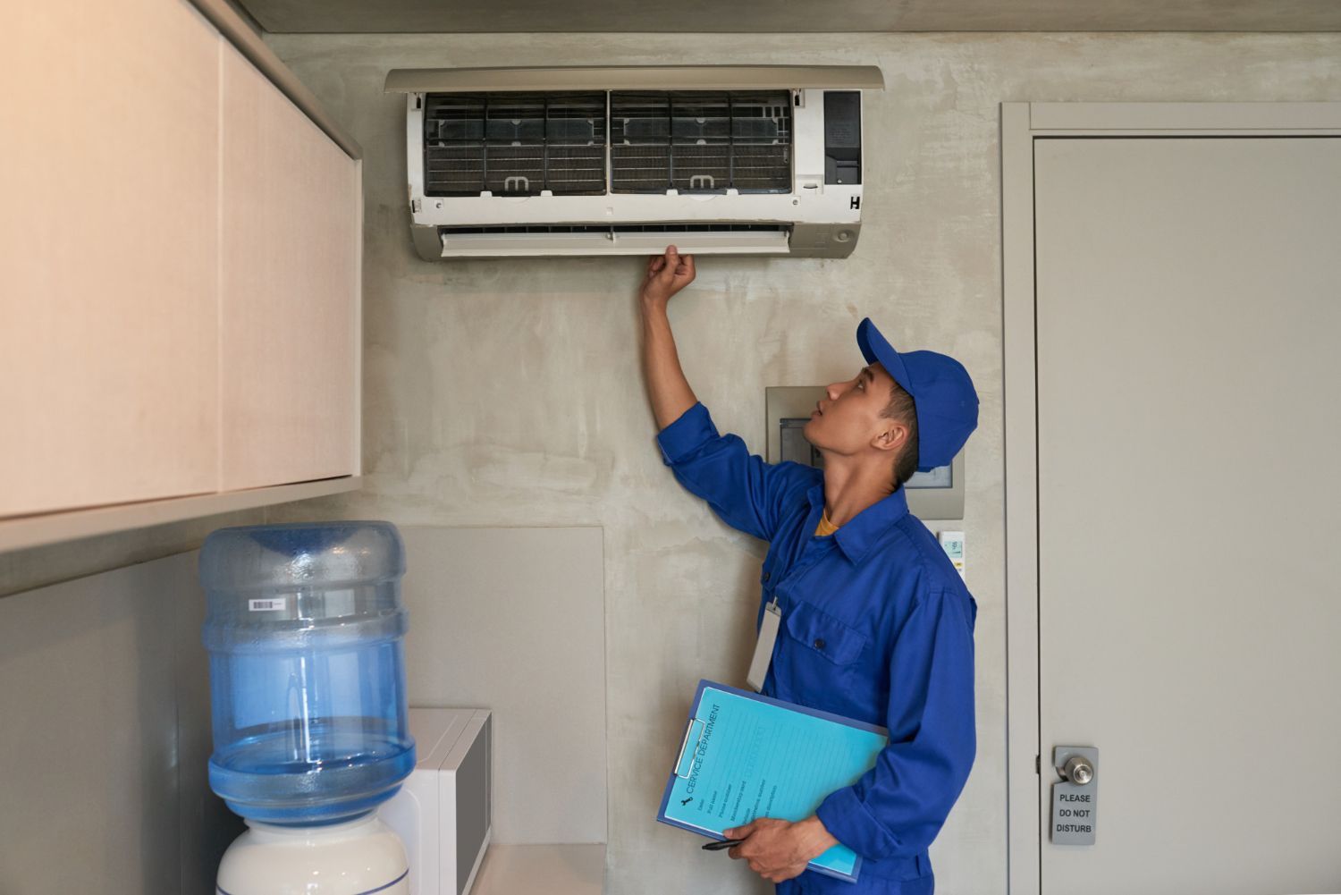 Technician in blue uniform inspecting a wall-mounted air conditioner indoors beside a water cooler.