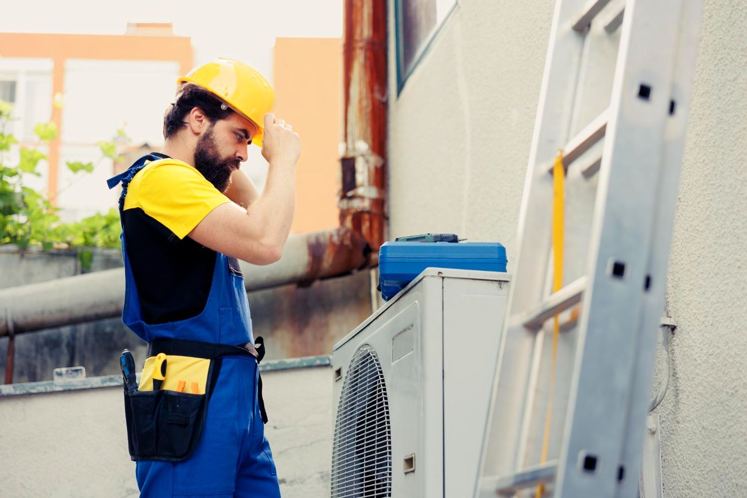 Technician in yellow hard hat inspecting an outdoor air conditioning unit beside a building