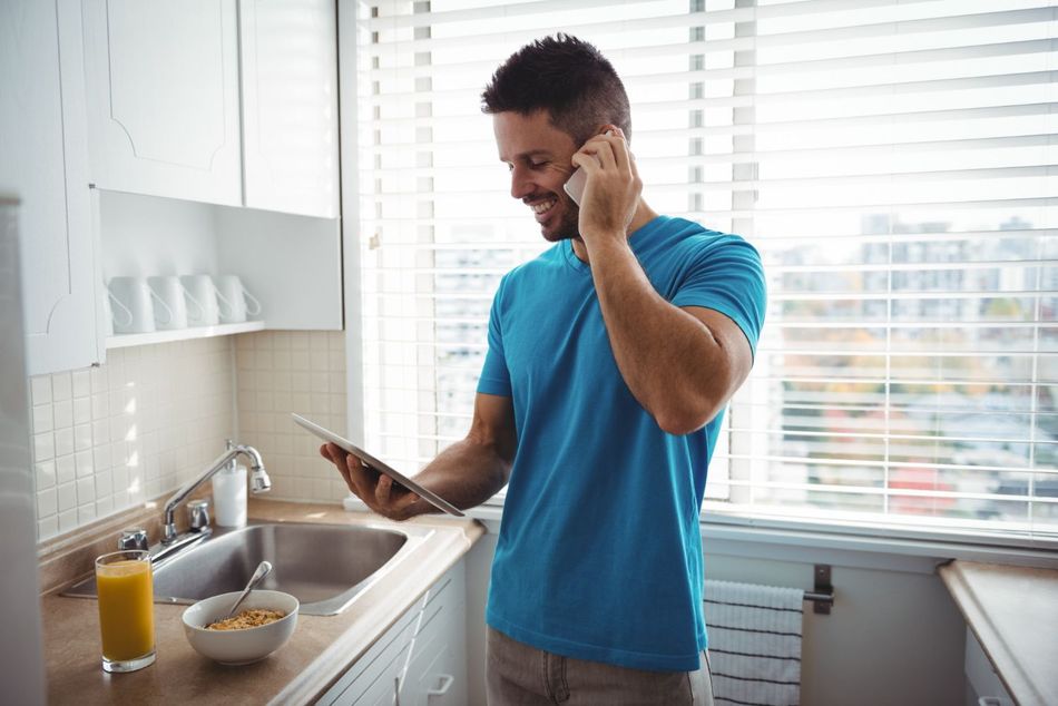 Man in a blue shirt talking on a phone in a bright kitchen while holding a smartphone near the sink.