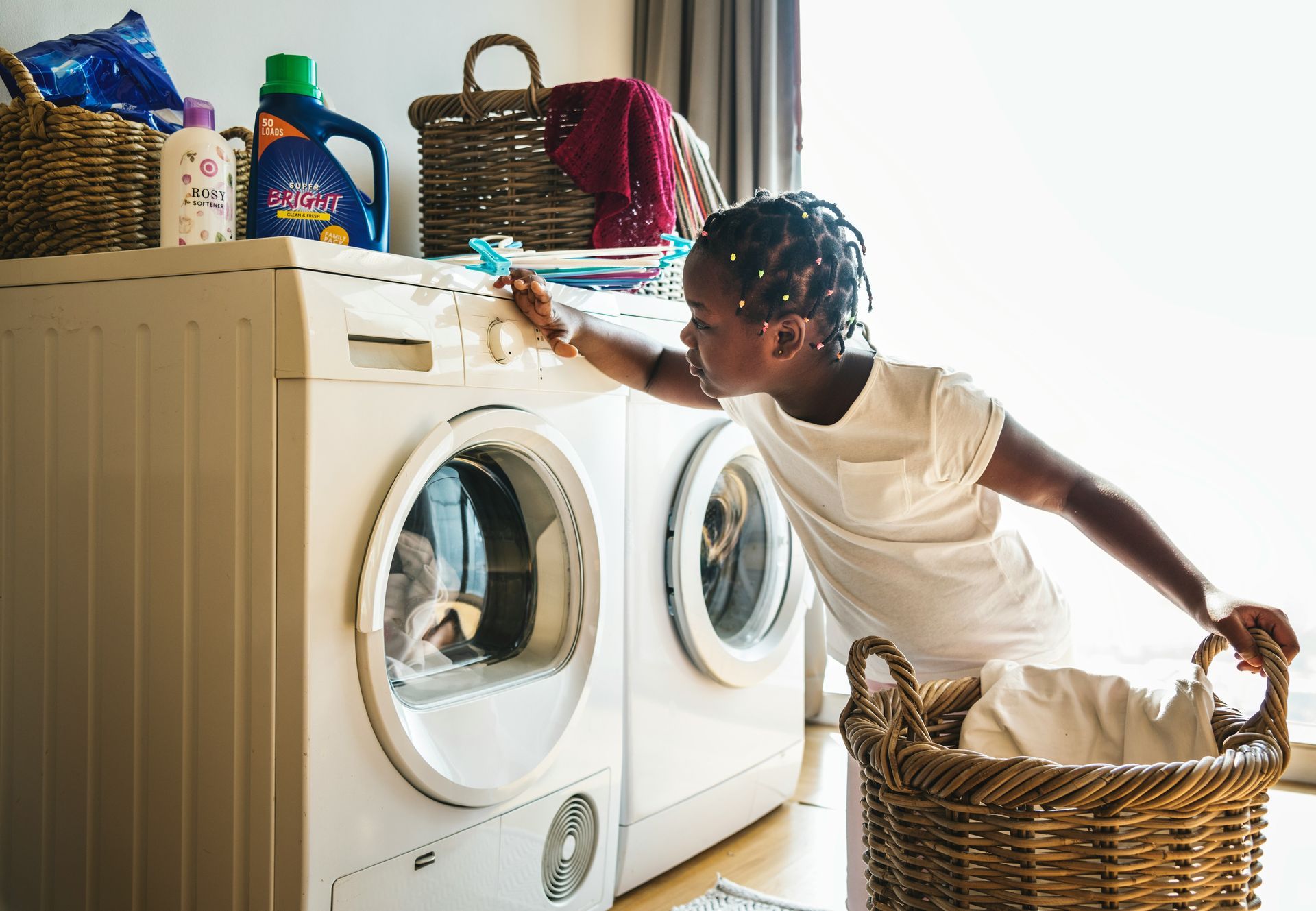 Woman loading laundry into a washing machine beside woven baskets and detergent bottles