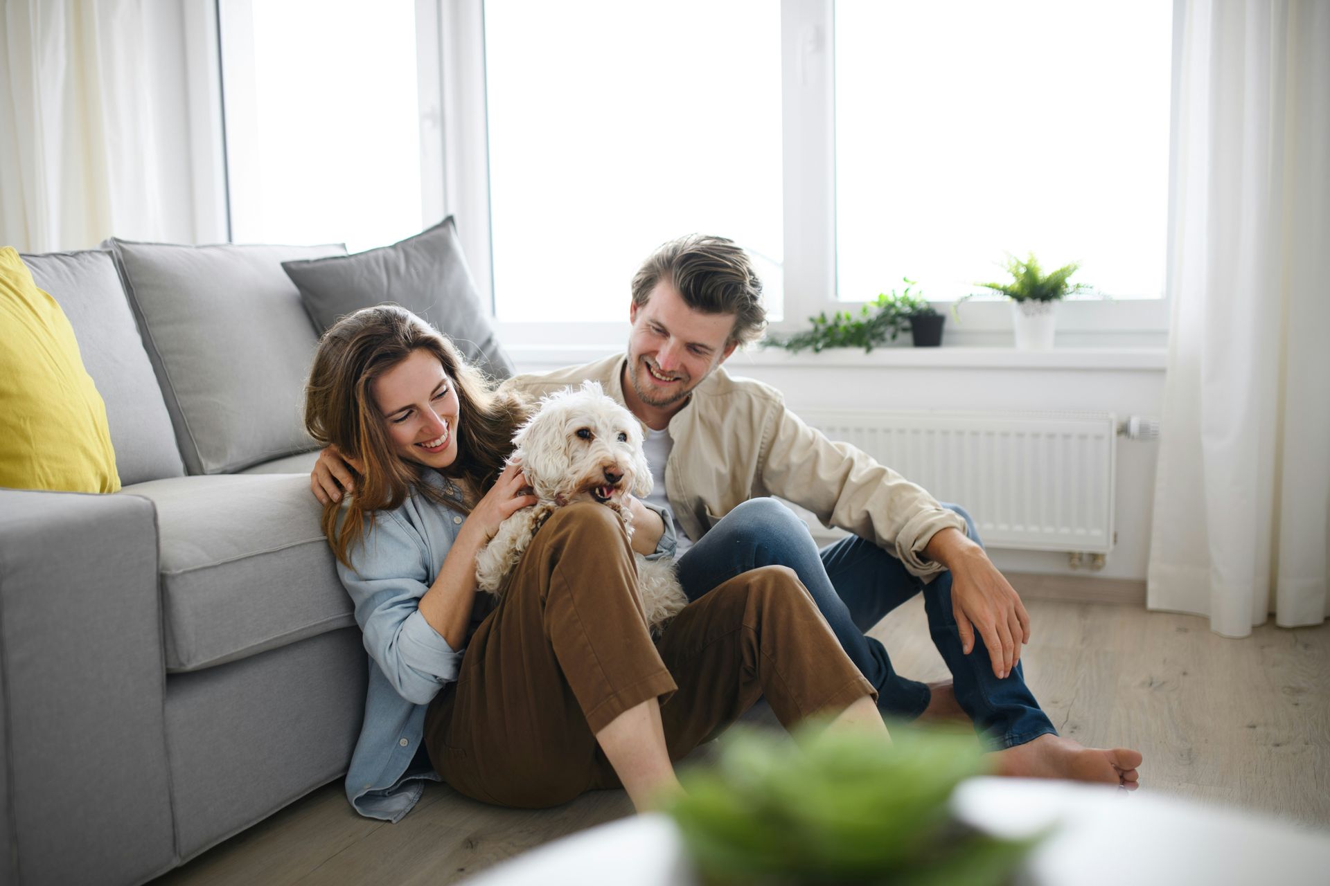 Couple sitting on a living room floor with a small white dog, smiling near a gray sofa and bright window