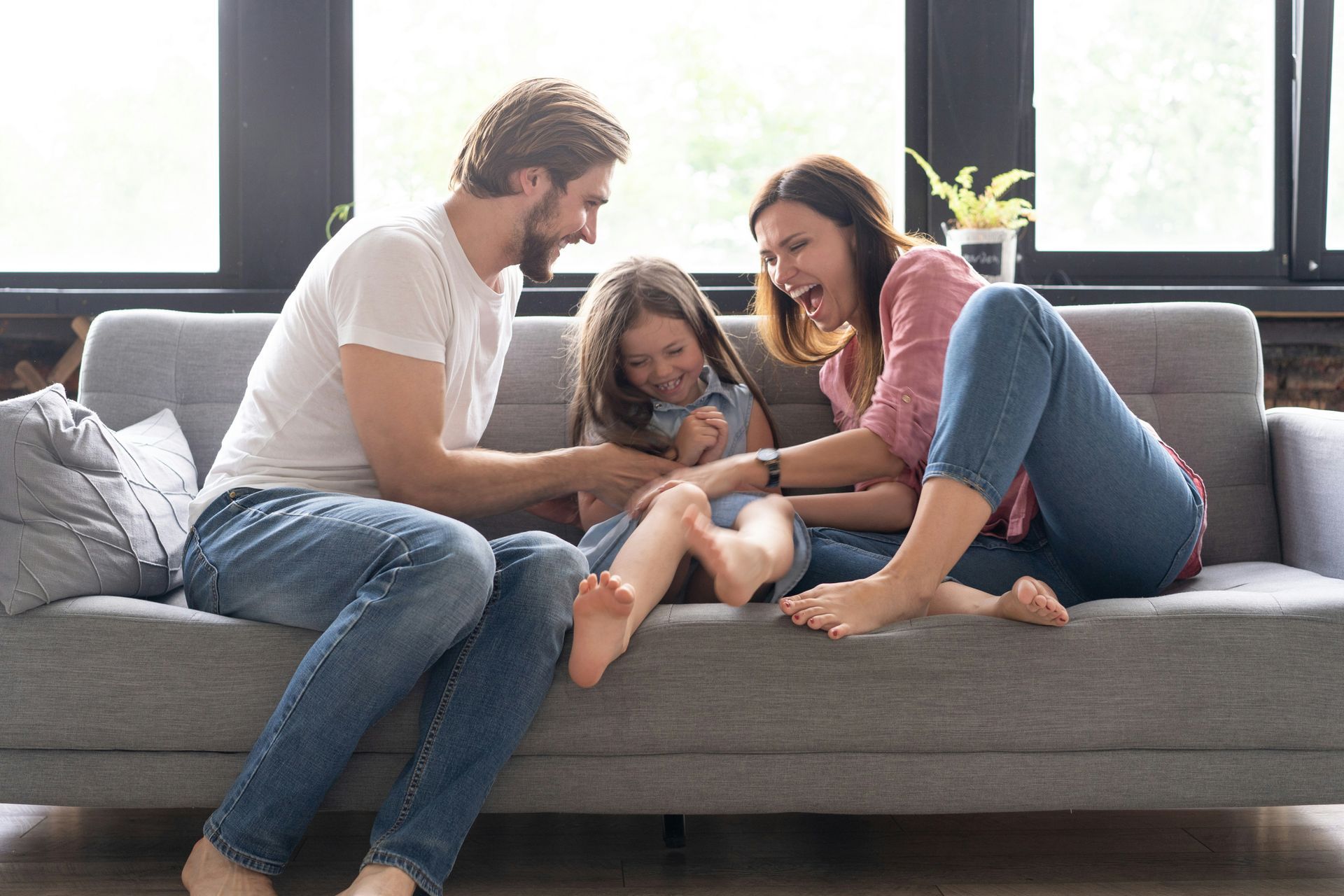 Family sitting on a gray couch, laughing and playing together in a bright living room.