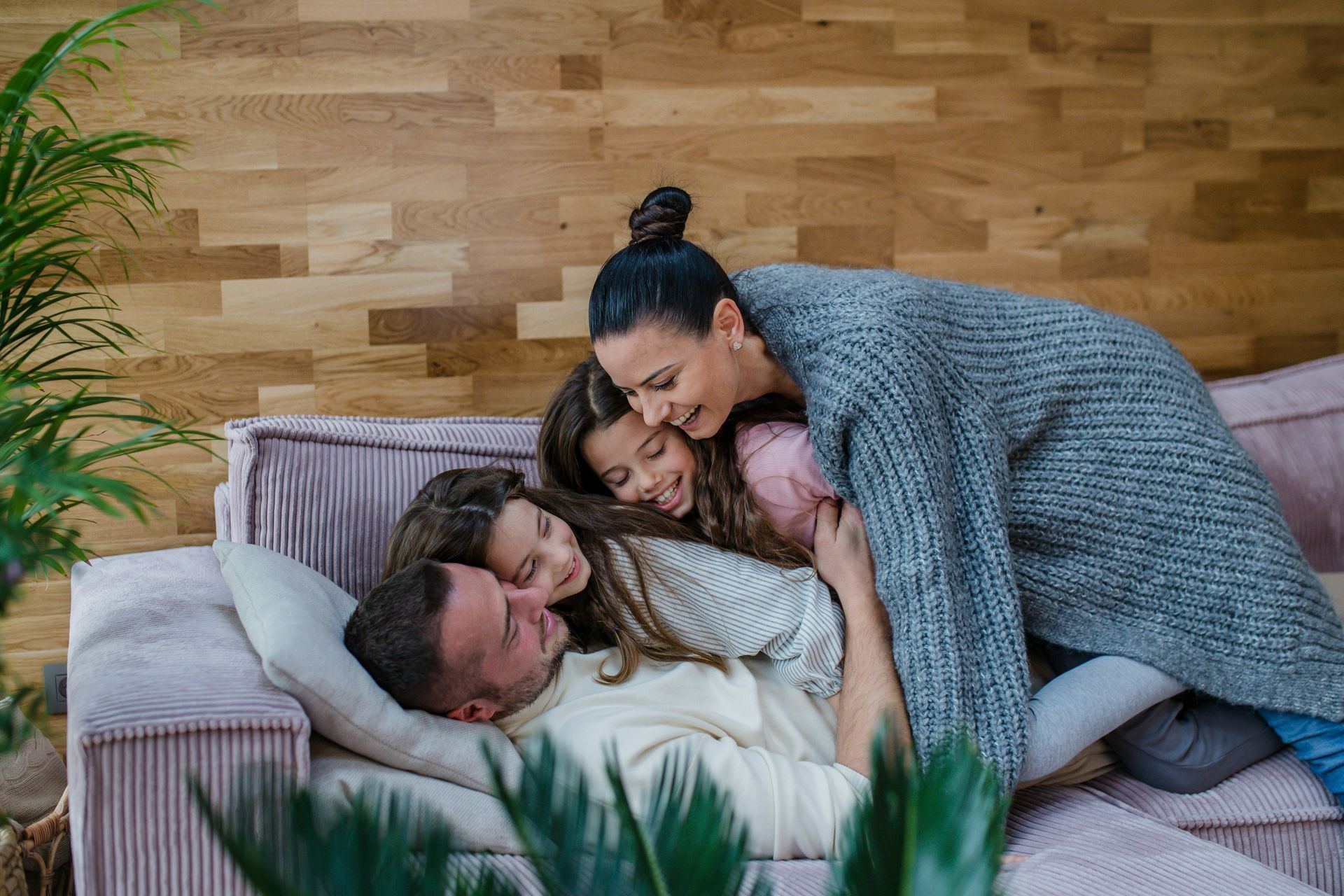 Three people cuddle on a couch in a cozy living room with wood-paneled walls.