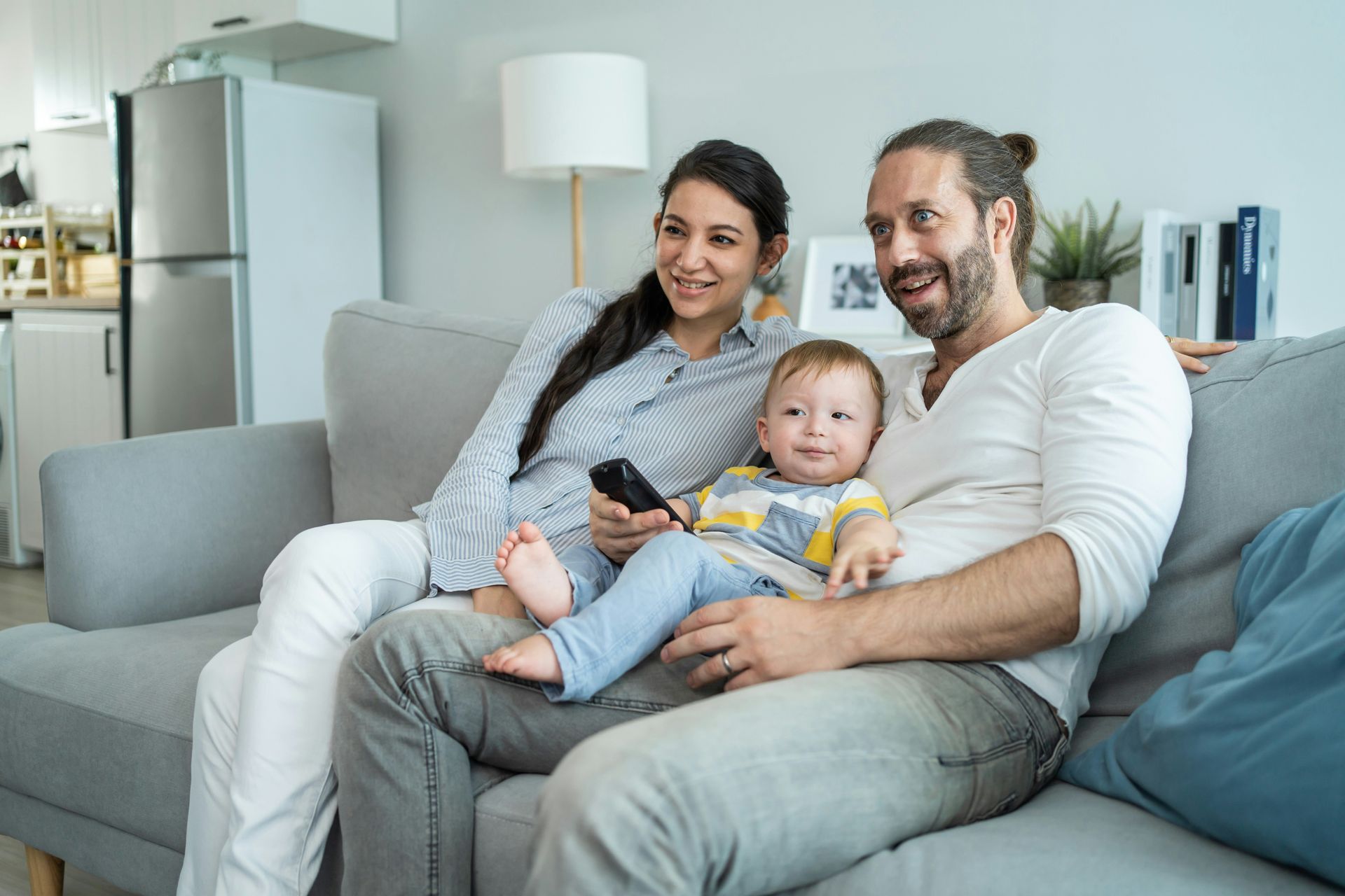 Family sitting on a gray couch, smiling and holding a baby in a bright living room