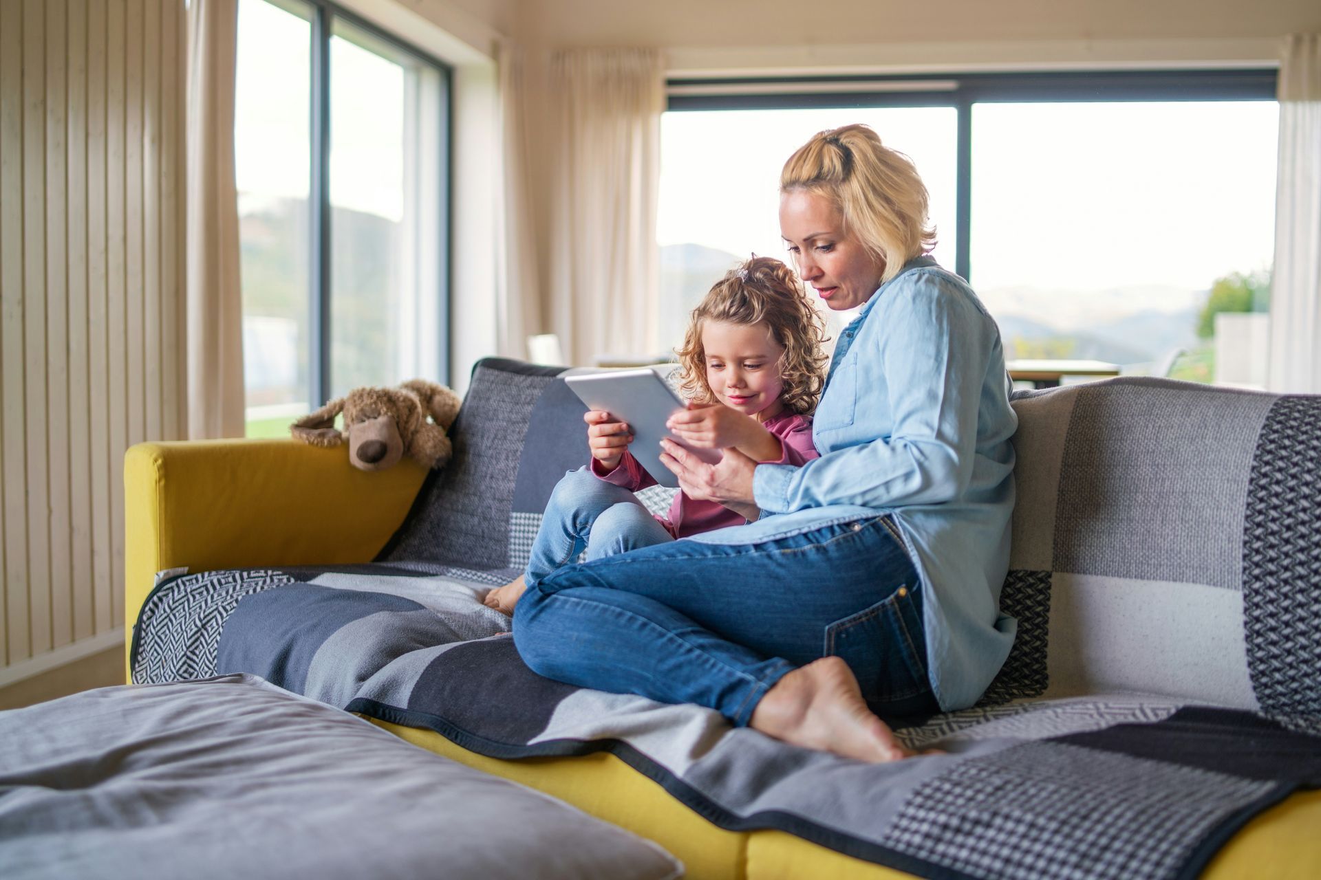 Woman and child sitting on a couch, looking at a tablet in a bright living room with a dog nearby