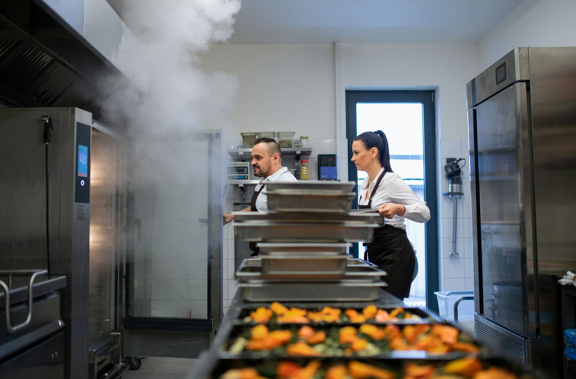 Commercial kitchen with chefs near steaming ovens and trays of food line-up on a counter