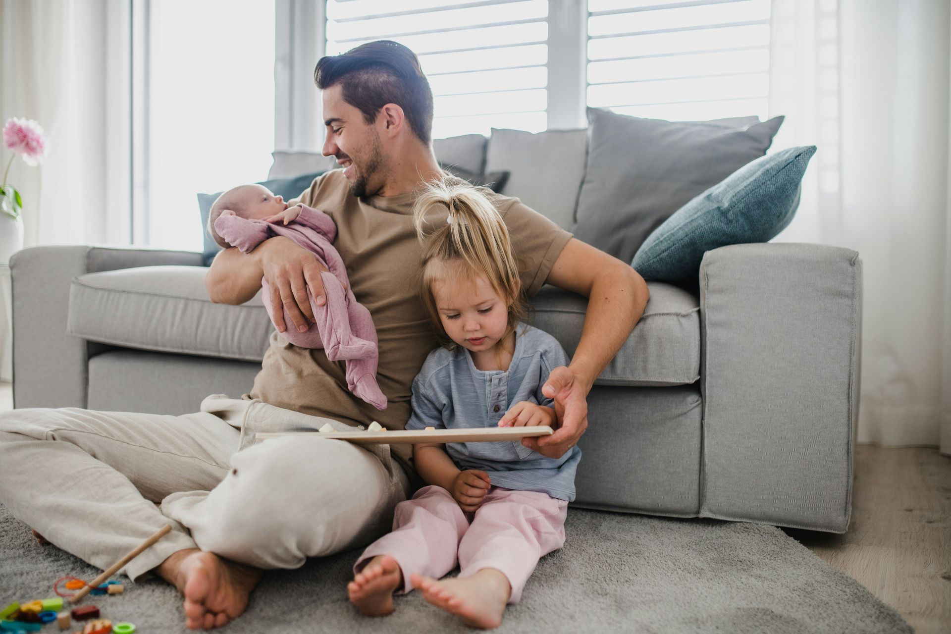 Parent sitting on carpet with two children in a bright living room, reading a book together.