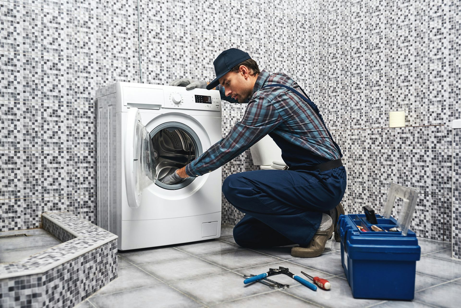 Technician repairing a front-loading washing machine beside tools and a blue bucket in a tiled laundry room