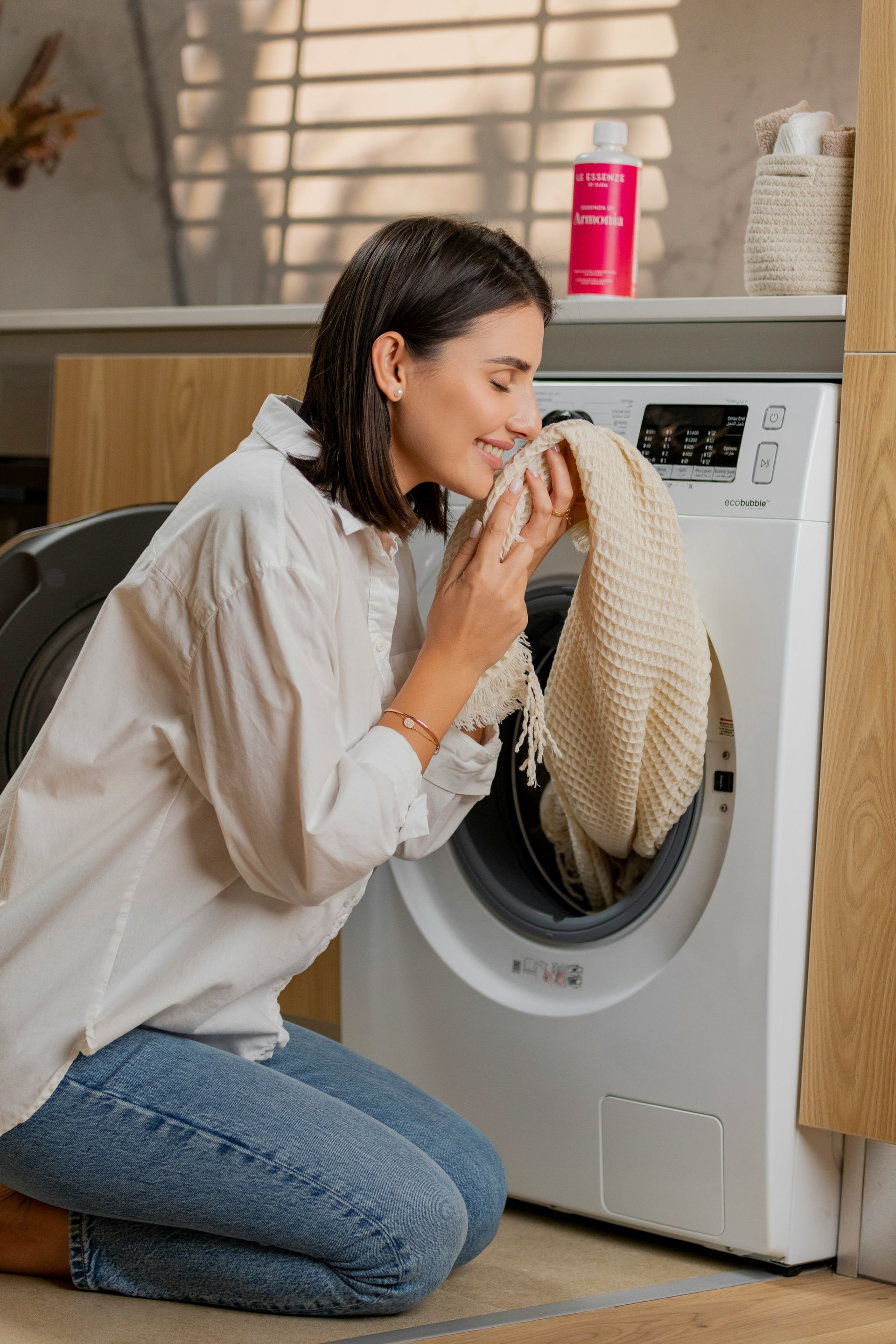 Woman kneeling by a washing machine, smiling as she smells a beige sweater in a bright laundry room.