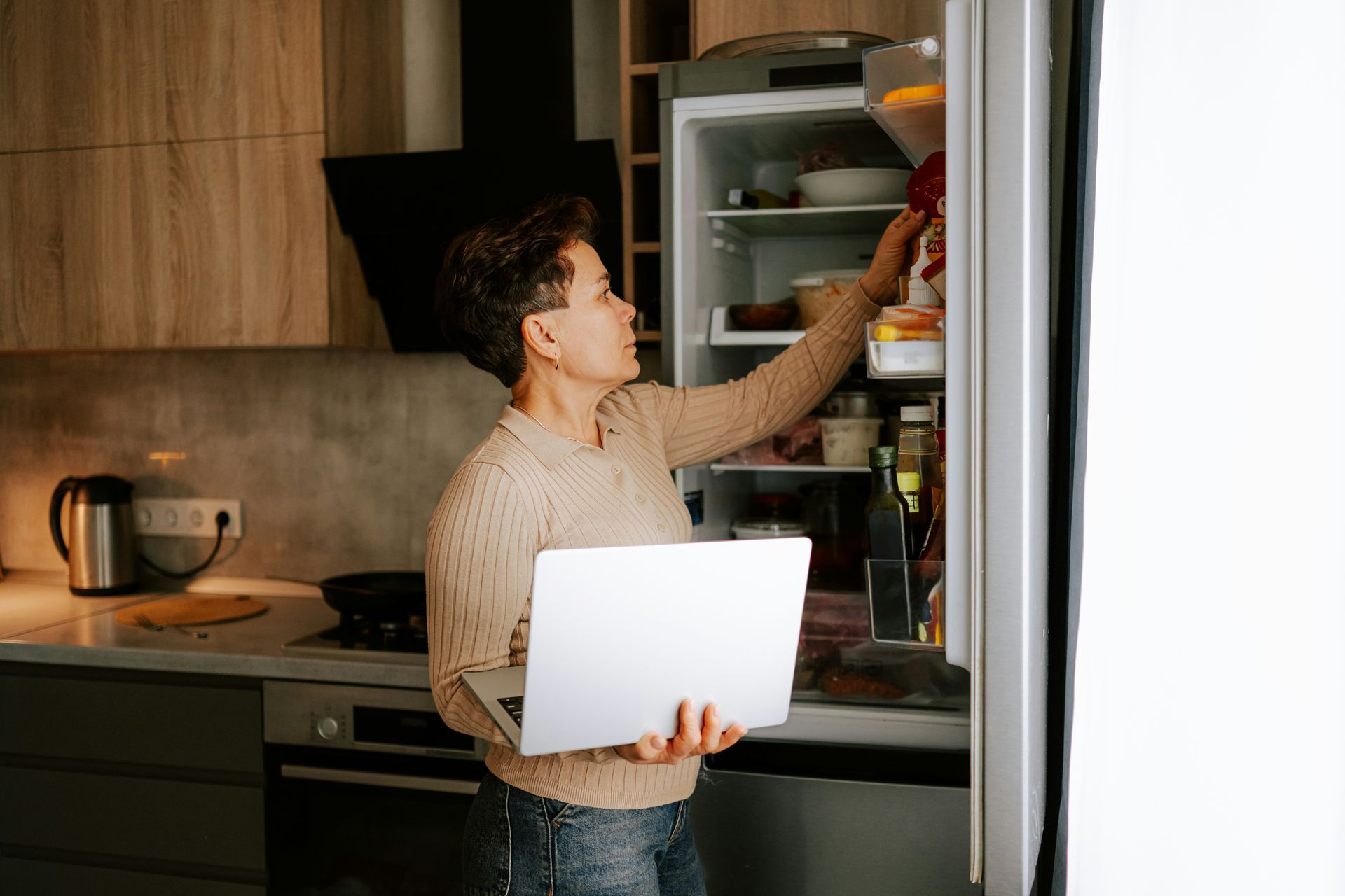 Person holding a laptop while reaching into an open refrigerator in a kitchen.