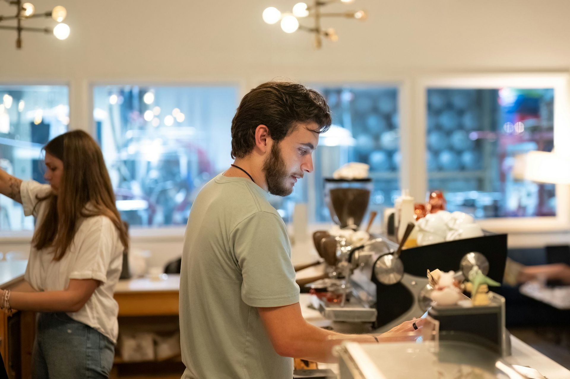 Person preparing coffee at a café counter while another customer stands in the background.