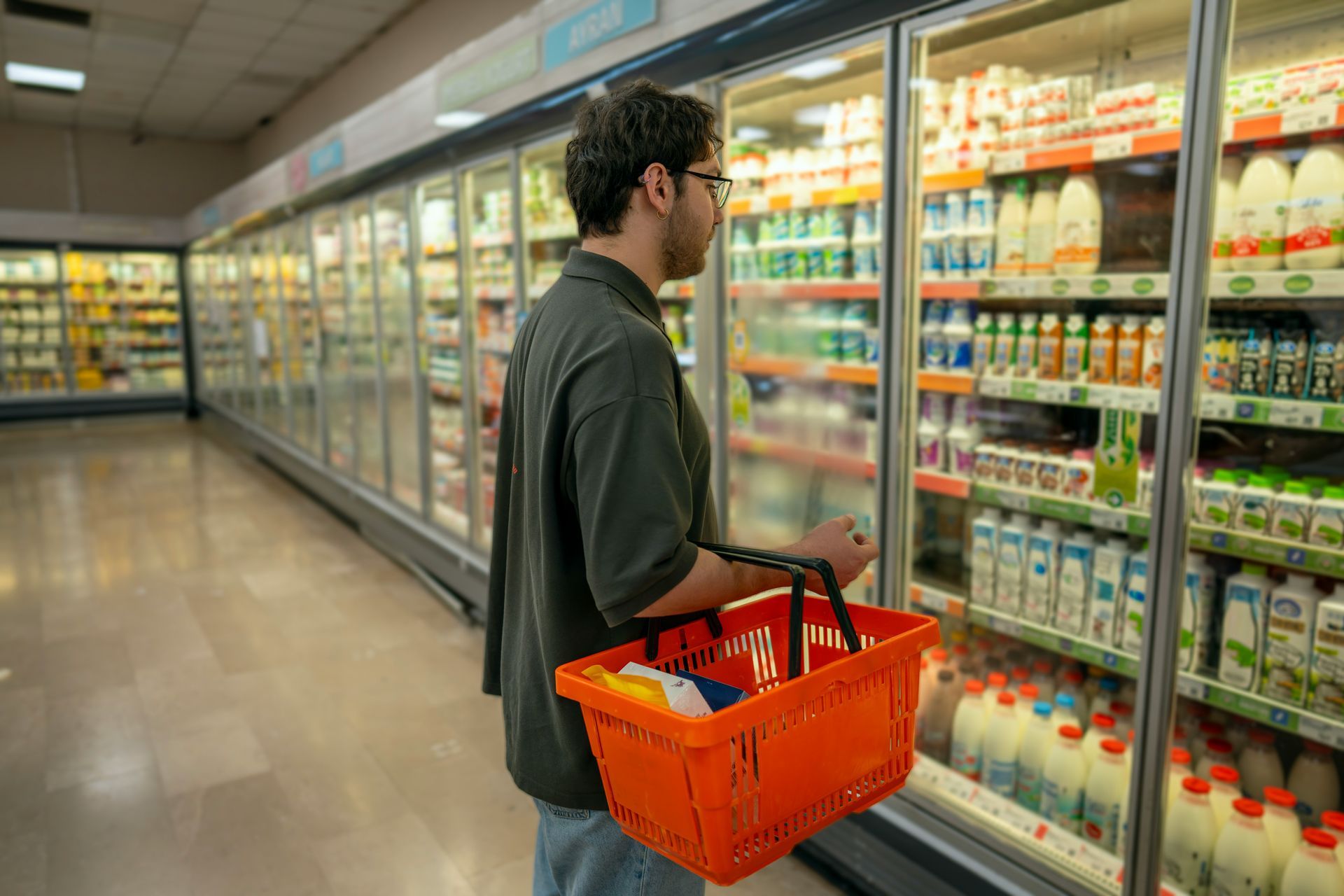 Person with an orange basket shopping in a brightly lit grocery store aisle by refrigerated shelves