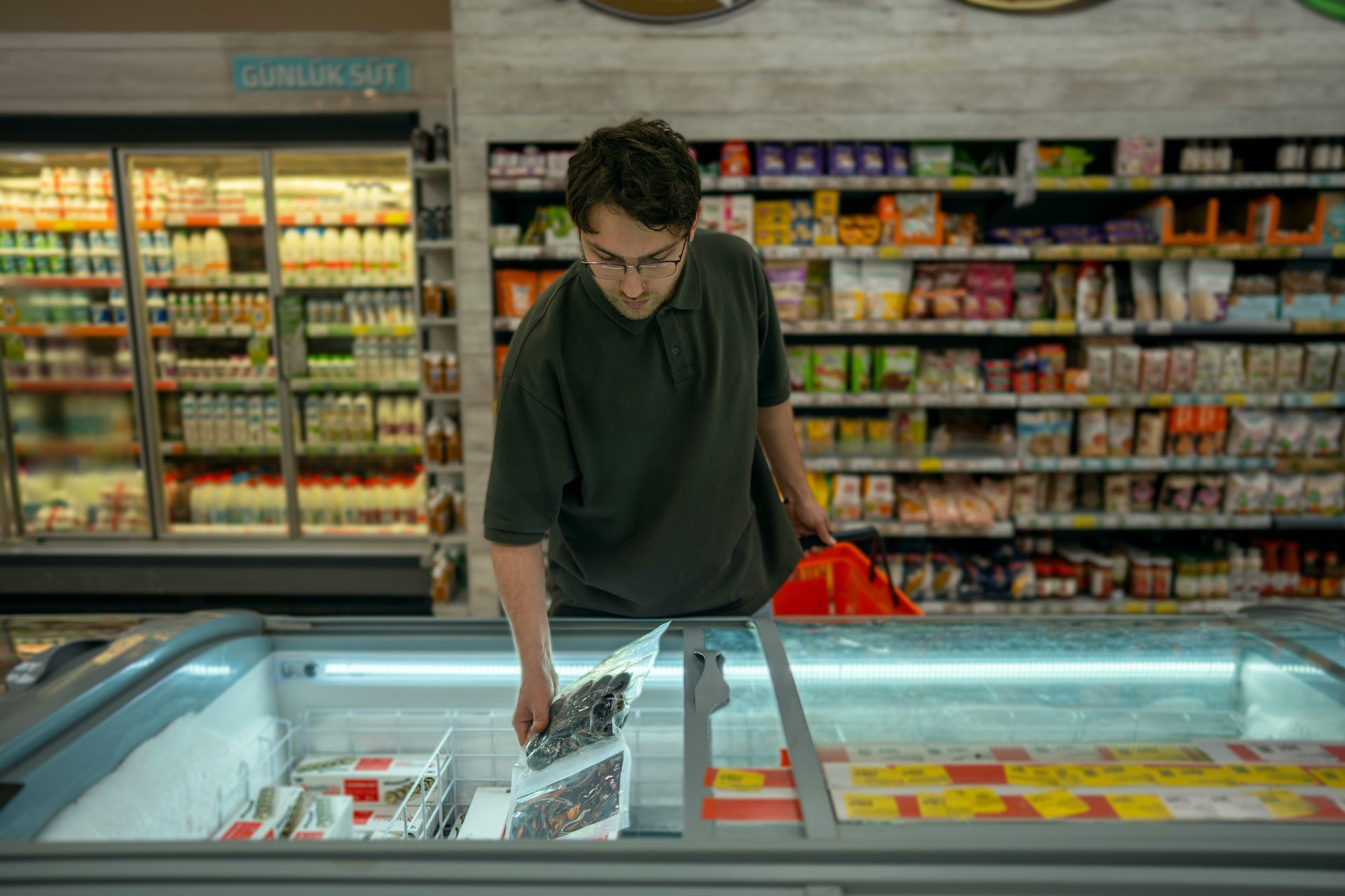 Man in a grocery freezer aisle selecting frozen items, with shelves of packaged goods behind him.