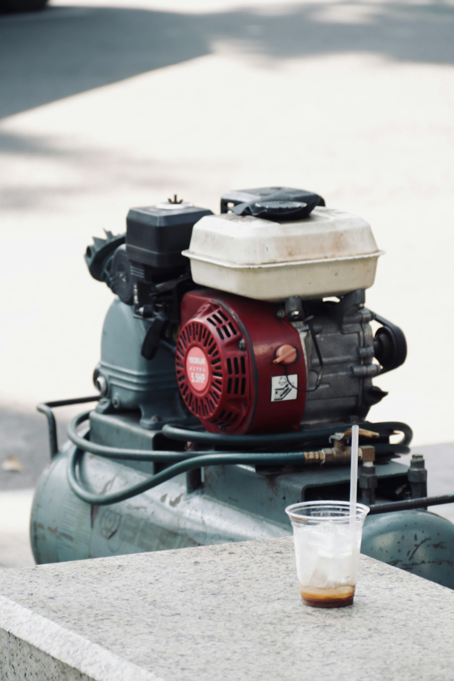 Small engine on a tank with a clear plastic cup of brown liquid in front, outdoors on pavement