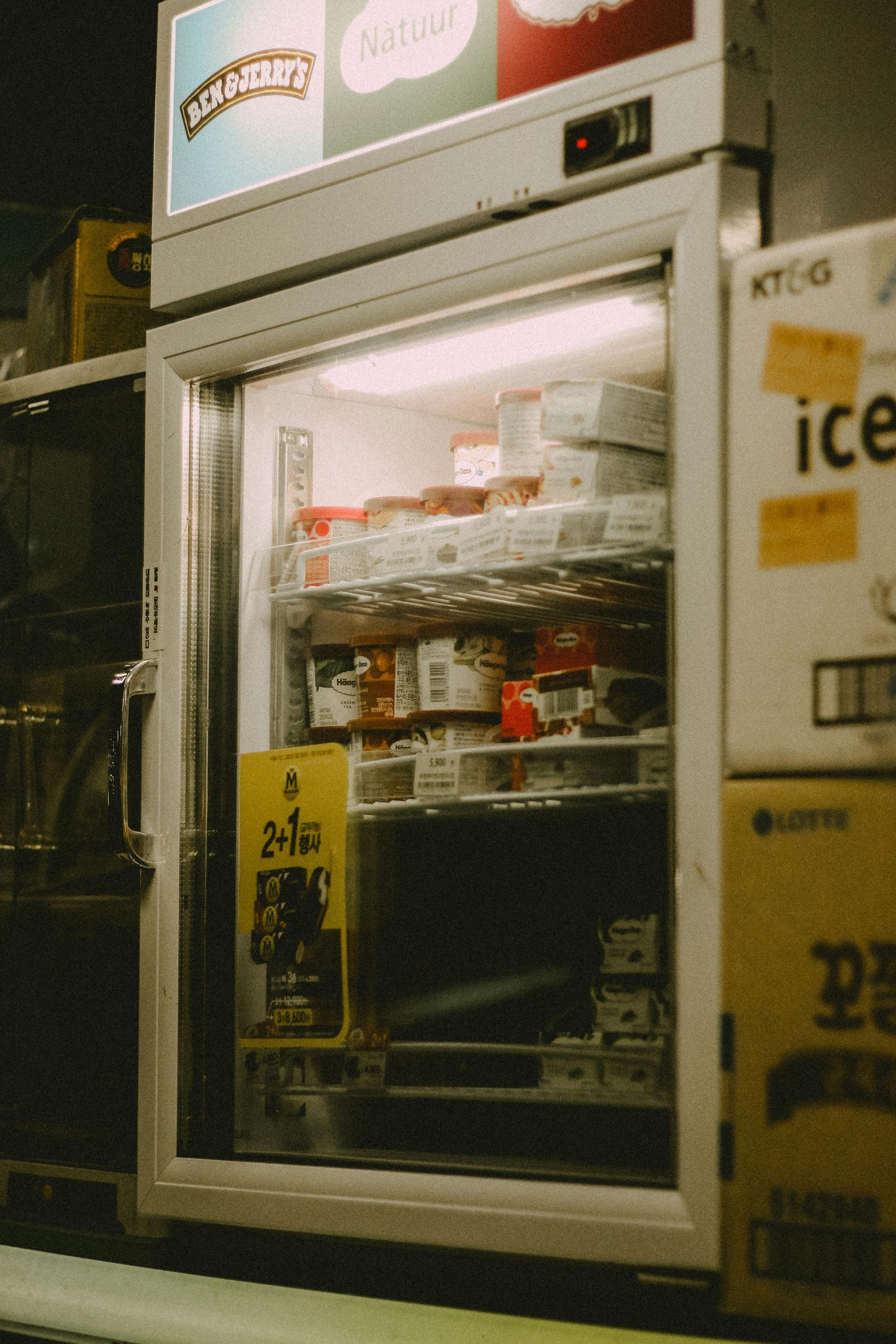 Dimly lit convenience store refrigerator stocked with drinks and snacks, with a white freezer door open on the right.