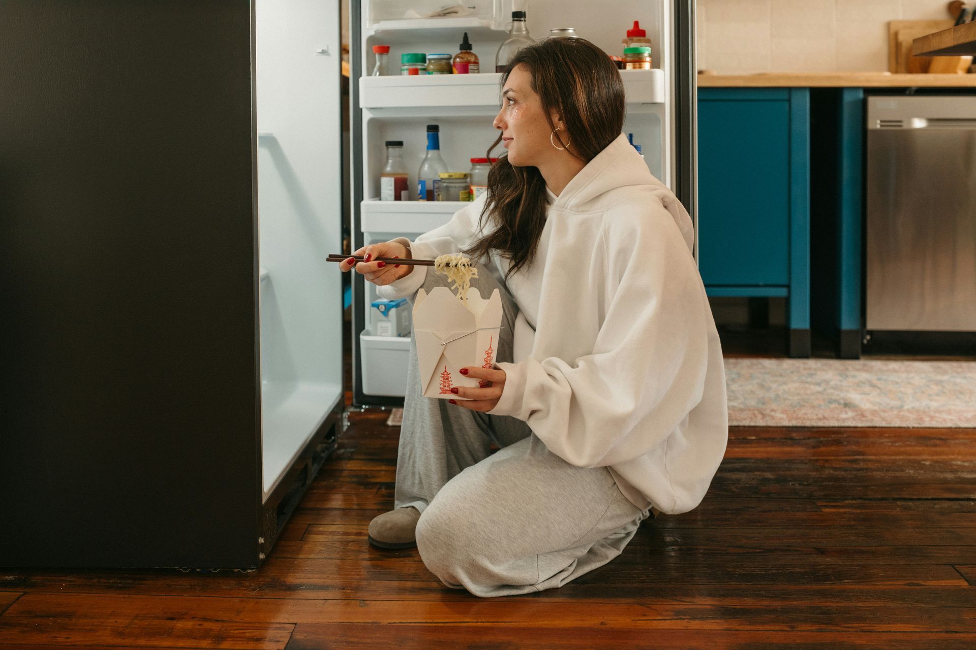 Woman crouching on a kitchen floor, holding a spray bottle and cleaning near a refrigerator.