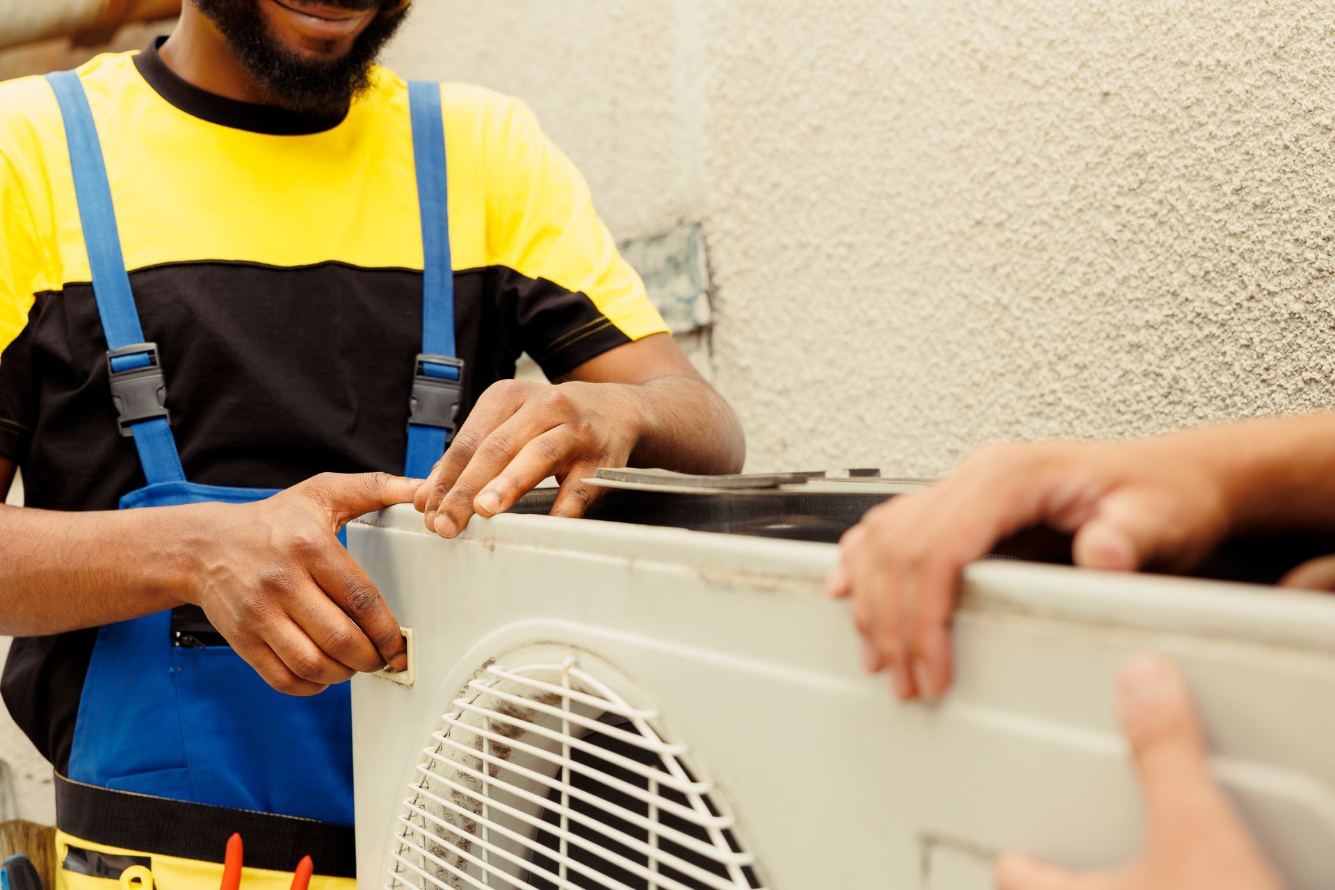 Technician in yellow shirt and blue overalls repairs an outdoor air conditioner unit while another person steadies it.