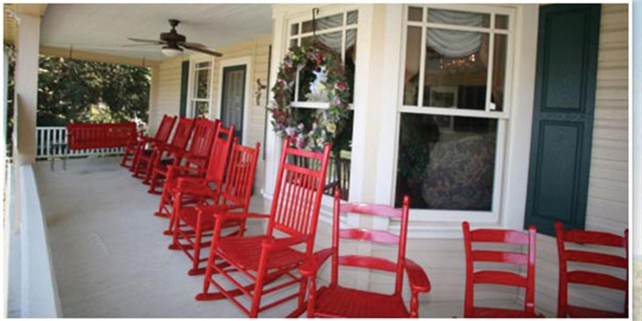 A long porch with red rocking chairs and a swing. A wreath hangs in the window.