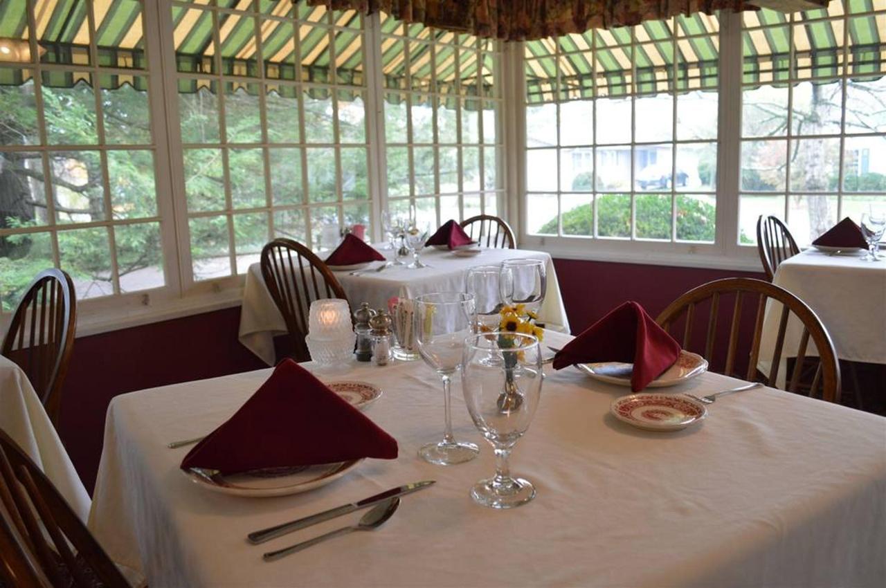 Elegant dining room with white tablecloths, red napkins, and wooden chairs, set near large windows overlooking a green outdoor area.