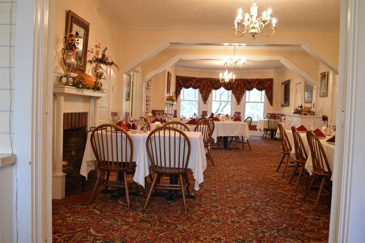 A dining room with tables set for a meal. Tables are covered with white cloths, wooden chairs surround them.