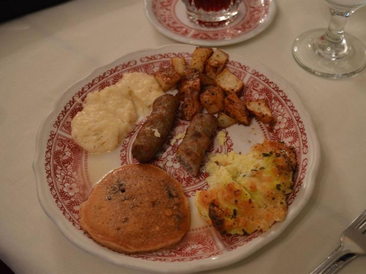 A plate of food with sausage, potatoes, a pancake, and other sides on a red-rimmed plate.