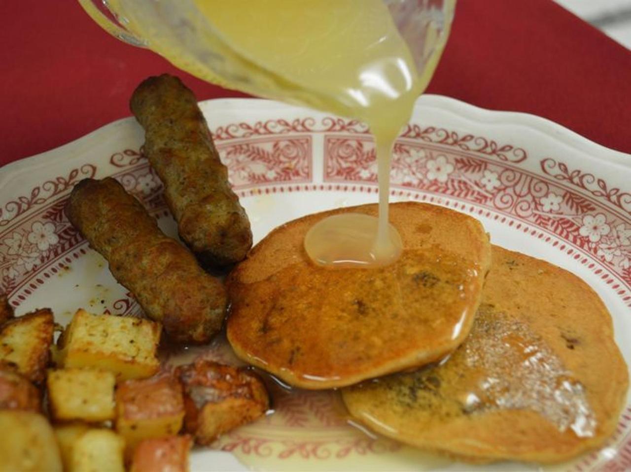 Breakfast plate with pancakes, sausage, and potatoes, being drizzled with syrup.
