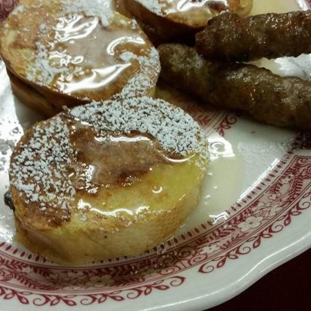 French toast with powdered sugar and syrup, served with sausage on a red-rimmed plate.